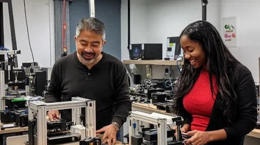 Two engineers assembling and testing black and silver 3D printers in a modern lab workspace