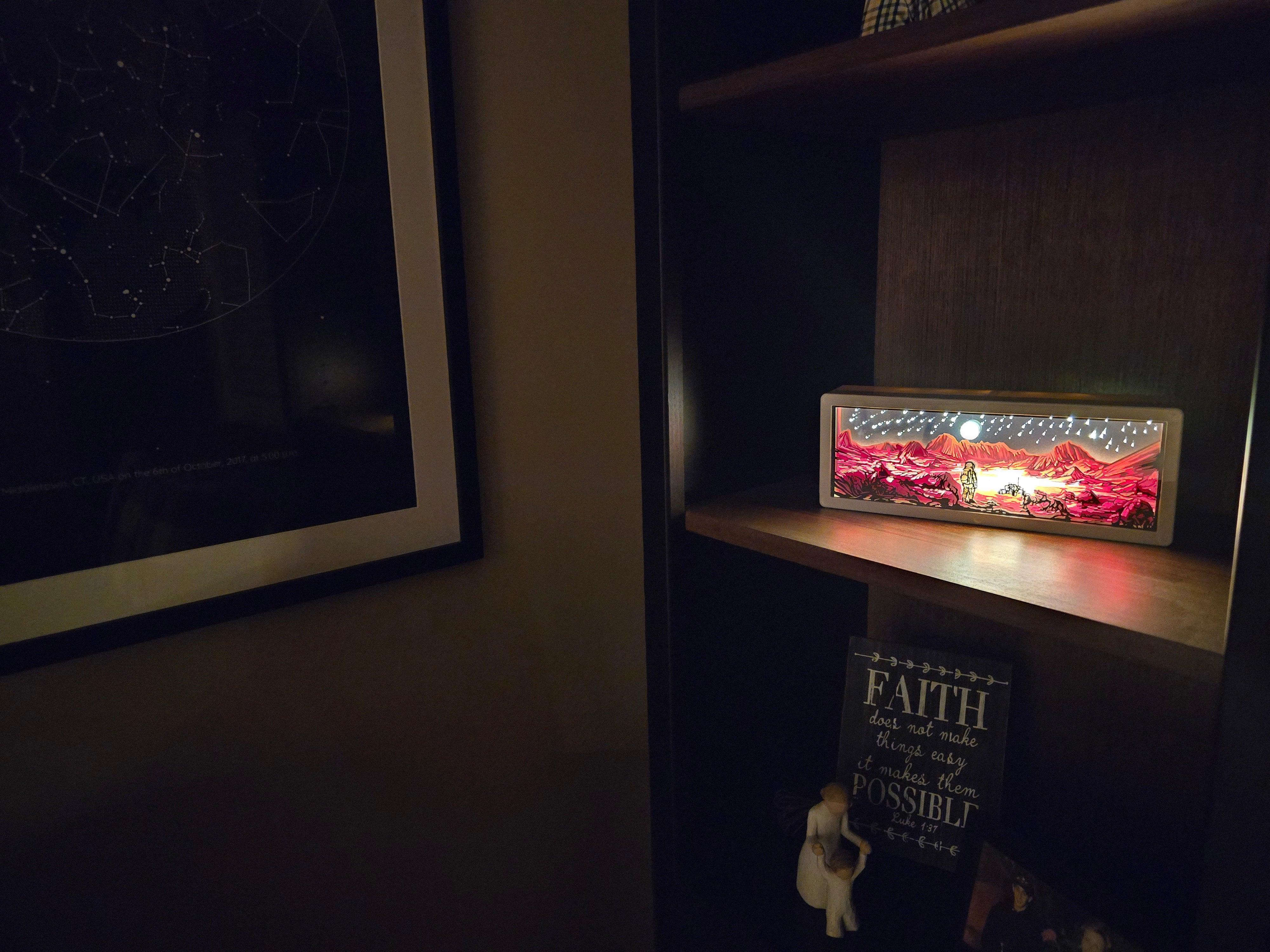 Dimly lit wooden shelf with a glowing framed artwork of a red alien landscape and starry sky