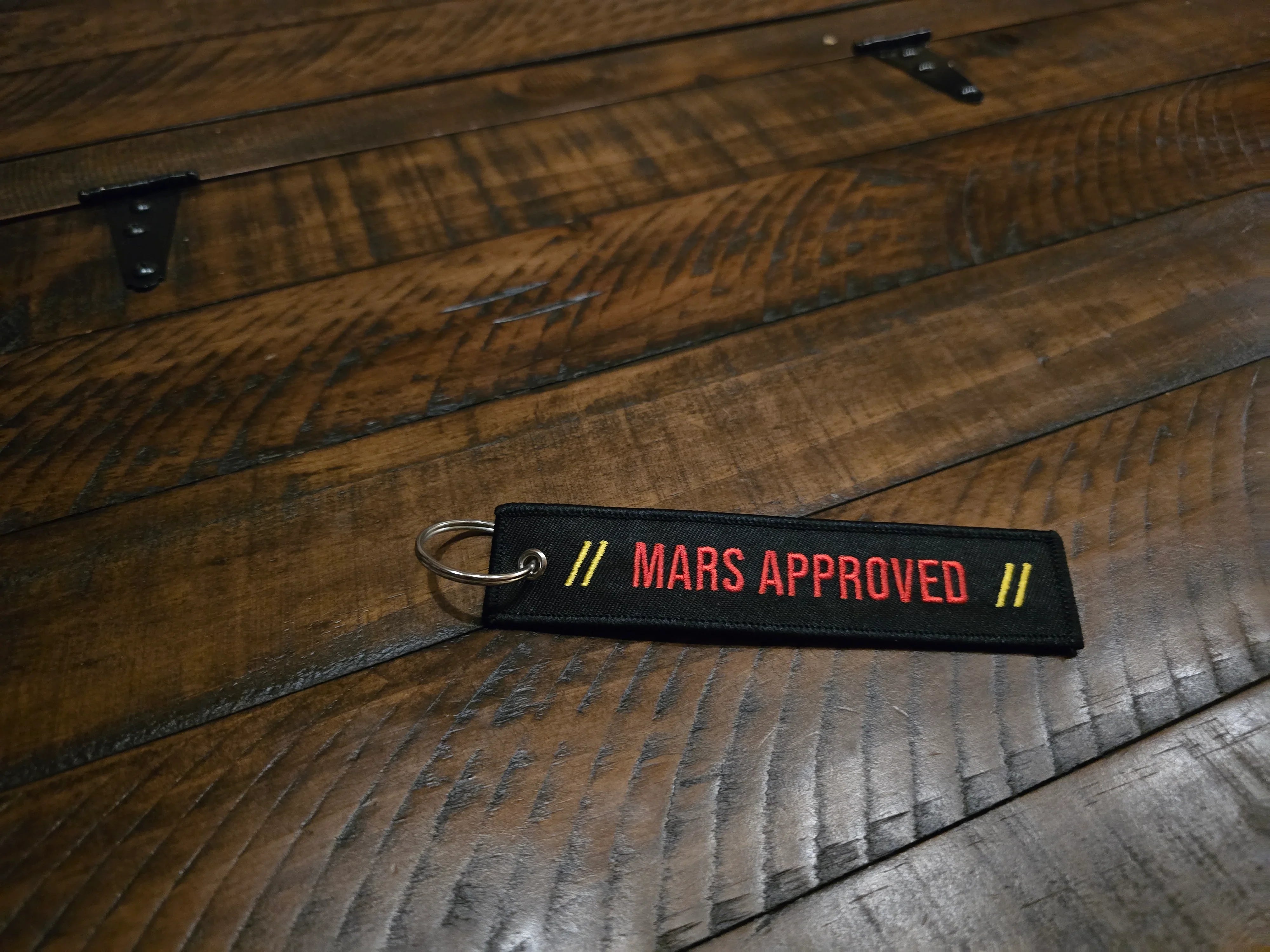 Black fabric keychain with red text 'MARS APPROVED' and yellow slashes, resting on rustic wooden surface