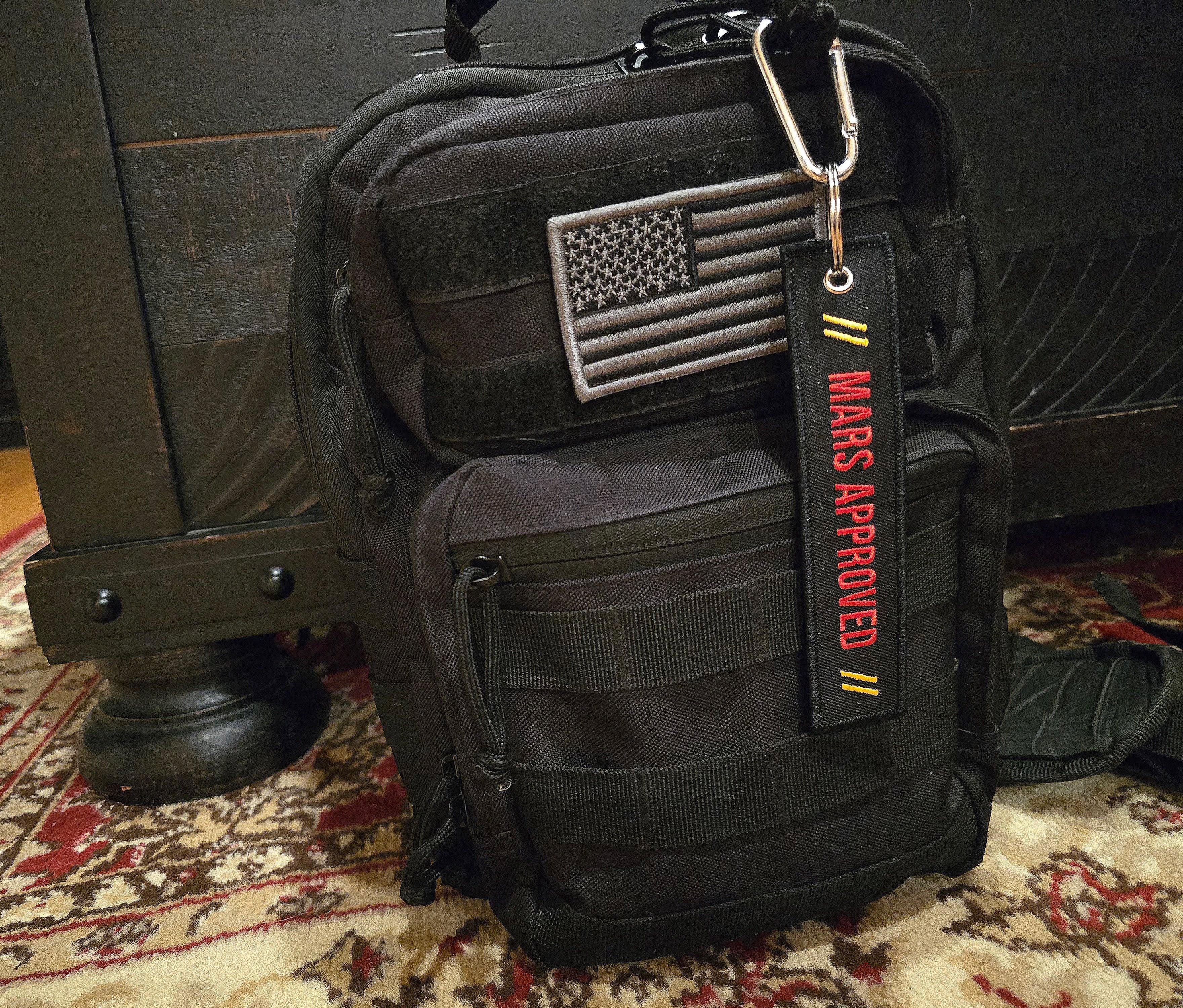 Black tactical backpack with American flag patch and "MARS APPROVED" keychain on a patterned carpet
