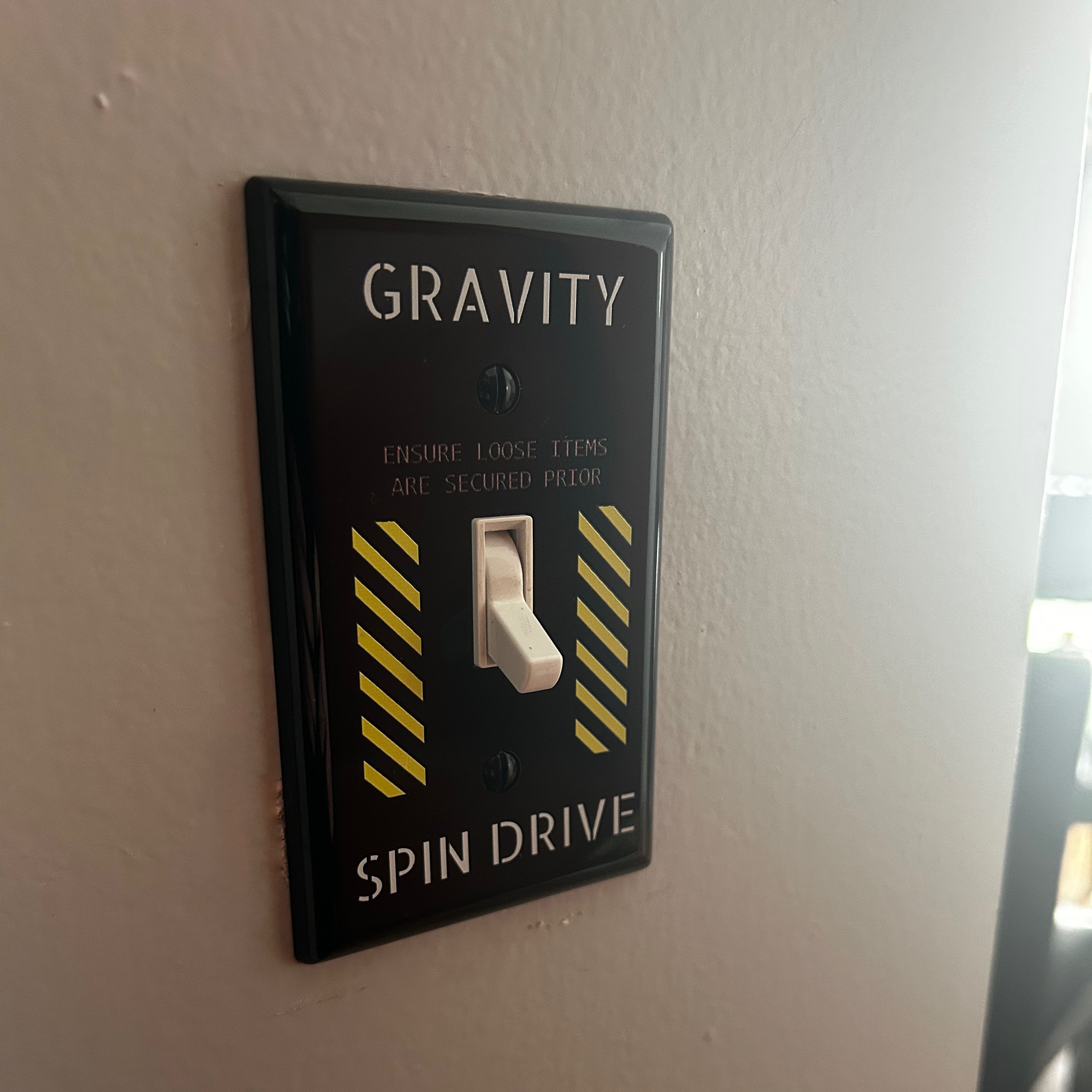 Decorative light switch cover with 'Gravity' and 'Spin Drive' text on a white wall.