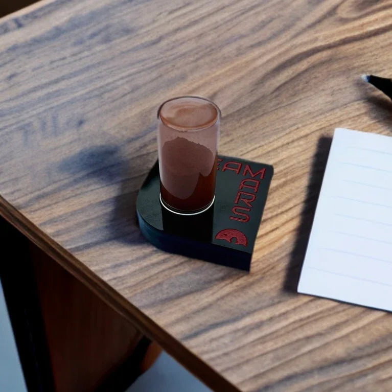 Shot glass with red liquid on black coaster labeled Team Mars on wooden table next to notepad and pen