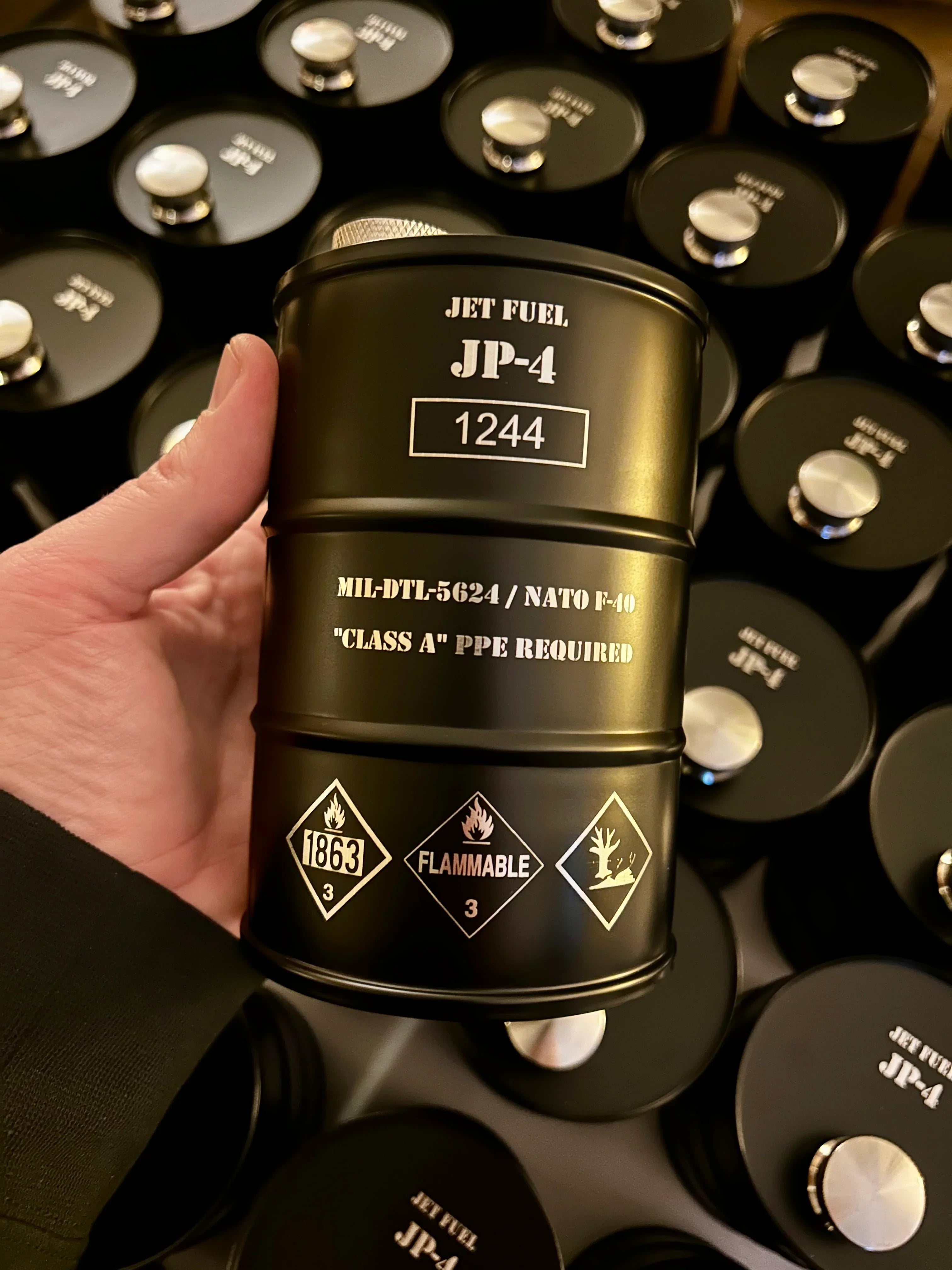 Hand holding a small black metal container labeled Jet Fuel JP-4 with hazard and flammable symbols, surrounded by similar containers