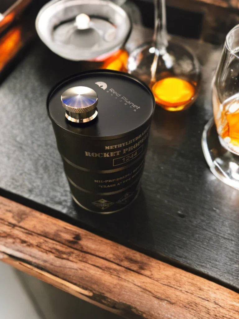 Black Red Planet Designs Rocket Propellant 1244 container with metal cap on dark table near whiskey glasses