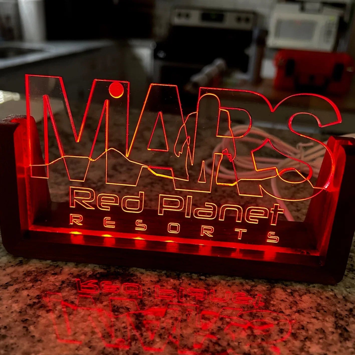 Glowing red acrylic sign on granite countertop reading 'Mars Red Planet Resorts' with mountain graphic