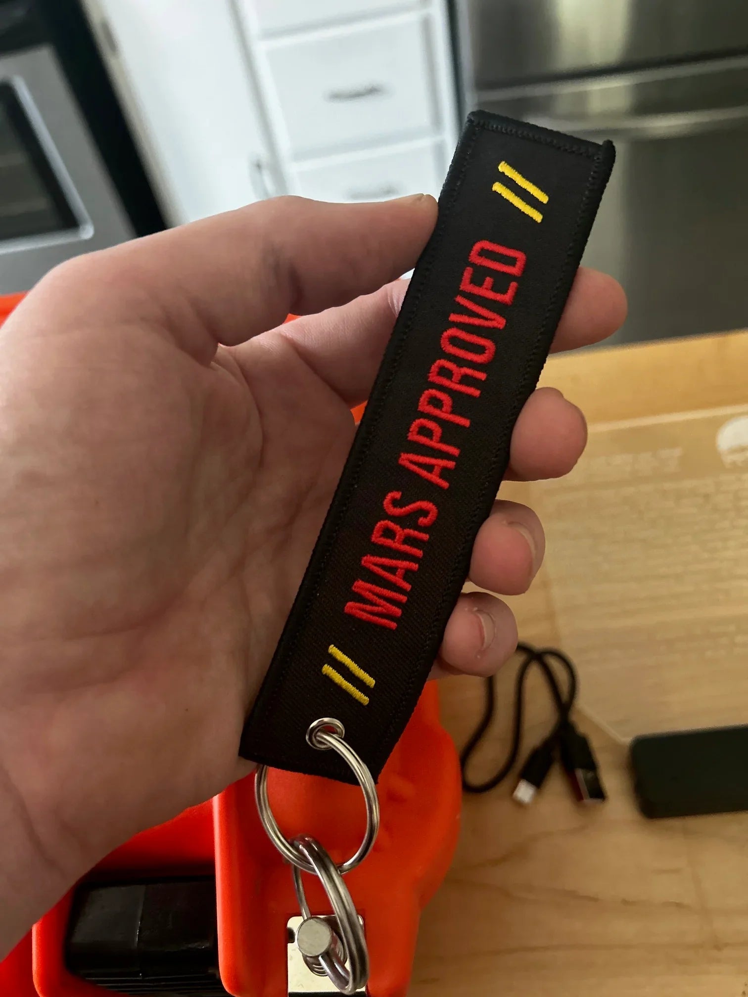 Hand holding black keychain with red text Mars Approved and yellow stripes, indoor setting