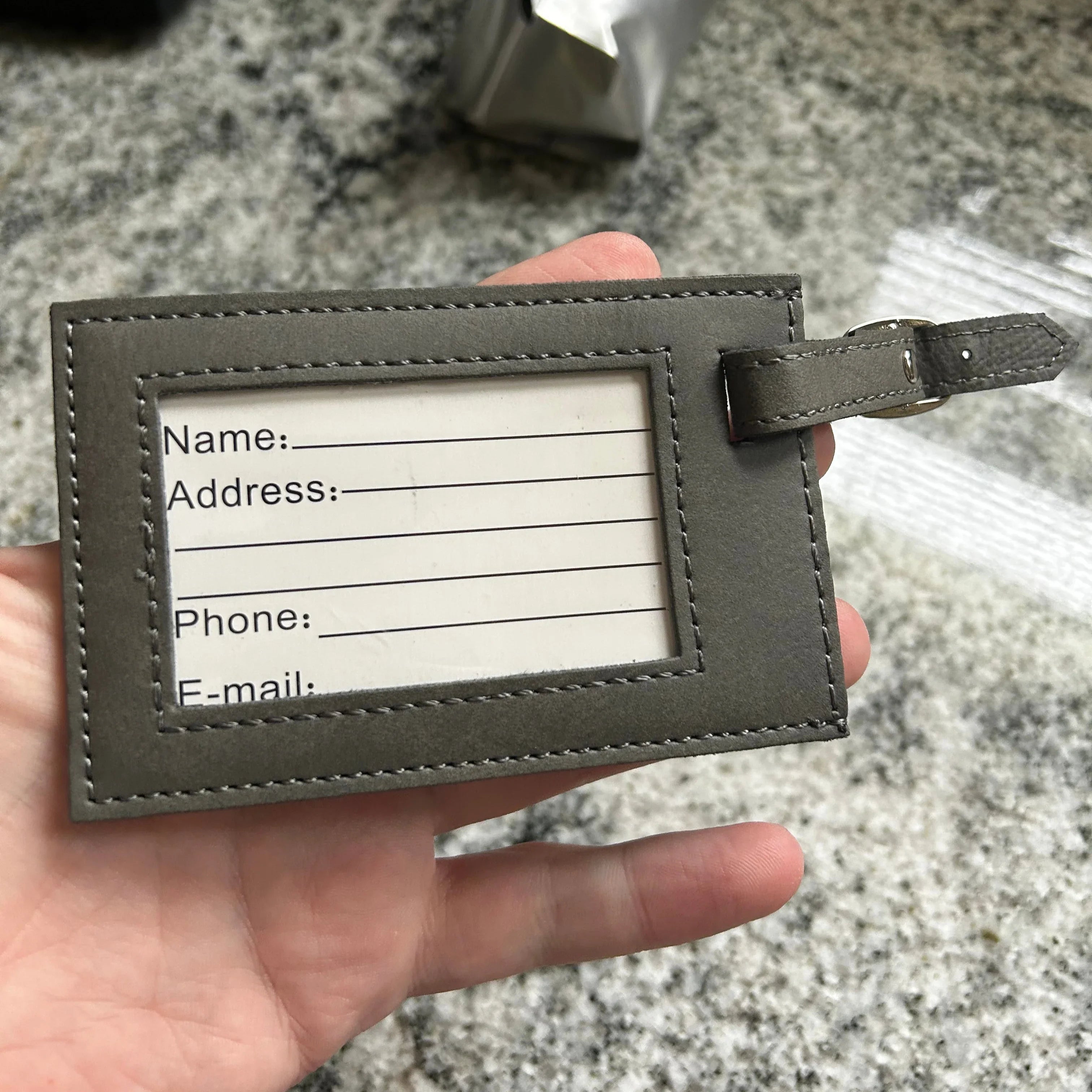 Hand holding a gray leather luggage tag with ID window and slots for name, address, phone, and email