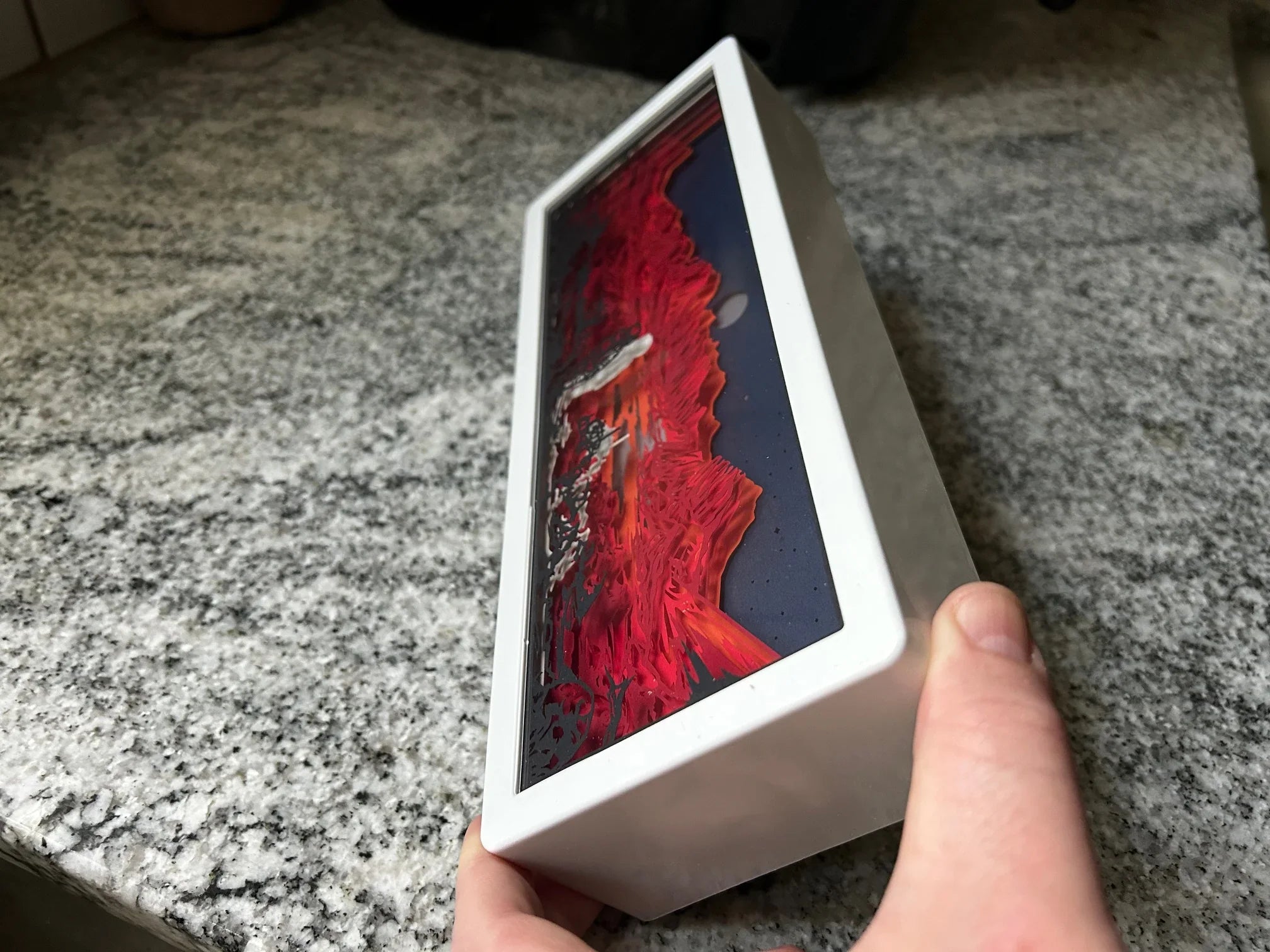 Hand holding a white box with red and black abstract design on top, on granite countertop