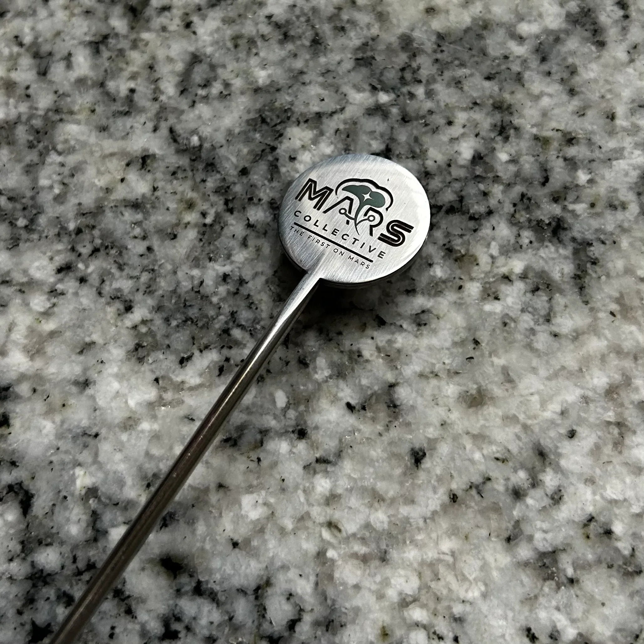 Metal spoon rest with Mars Collective logo and text on granite countertop