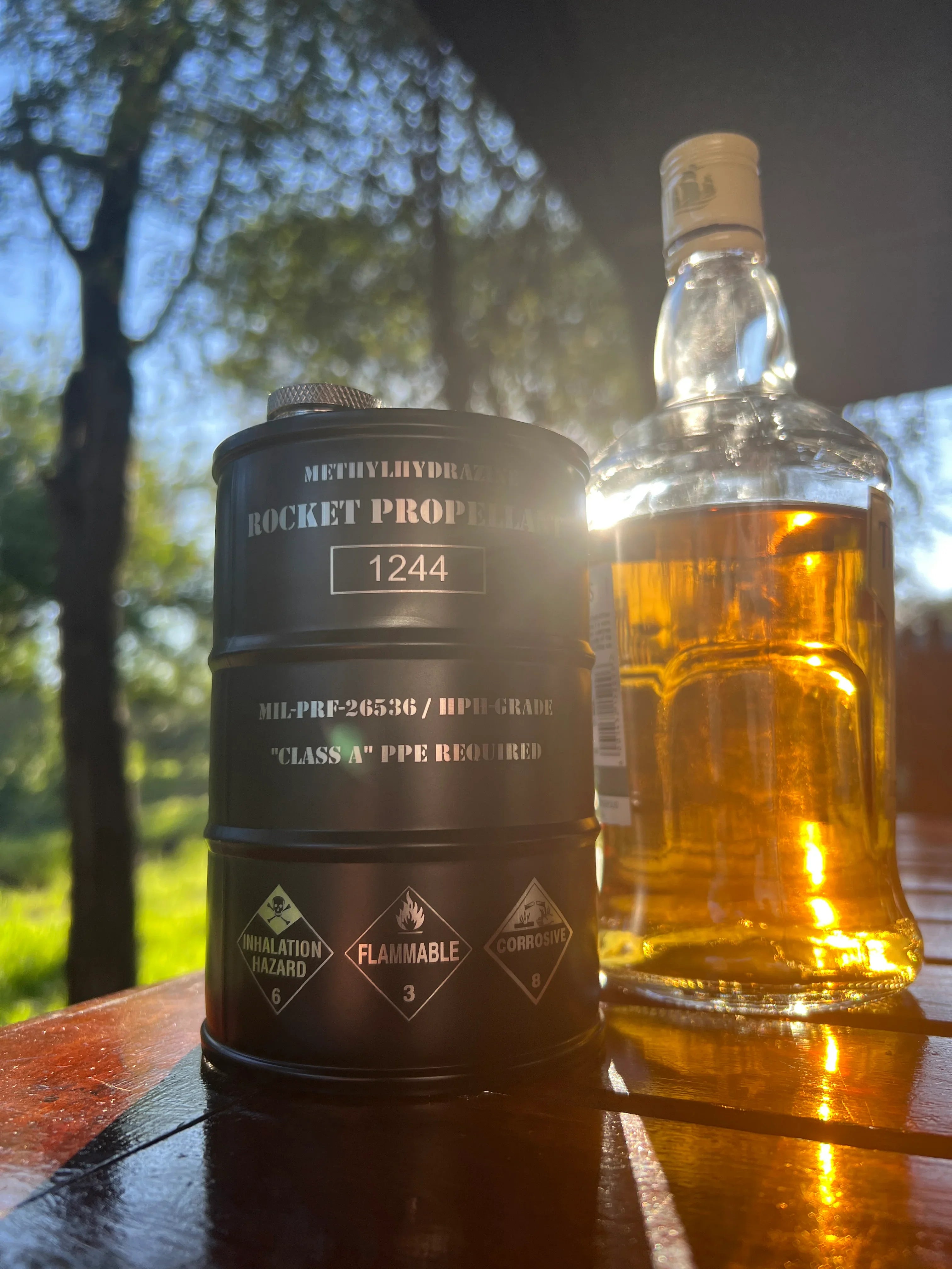 Black container labeled rocket propellant with hazard symbols next to a glass bottle of amber liquid on wooden table outdoors