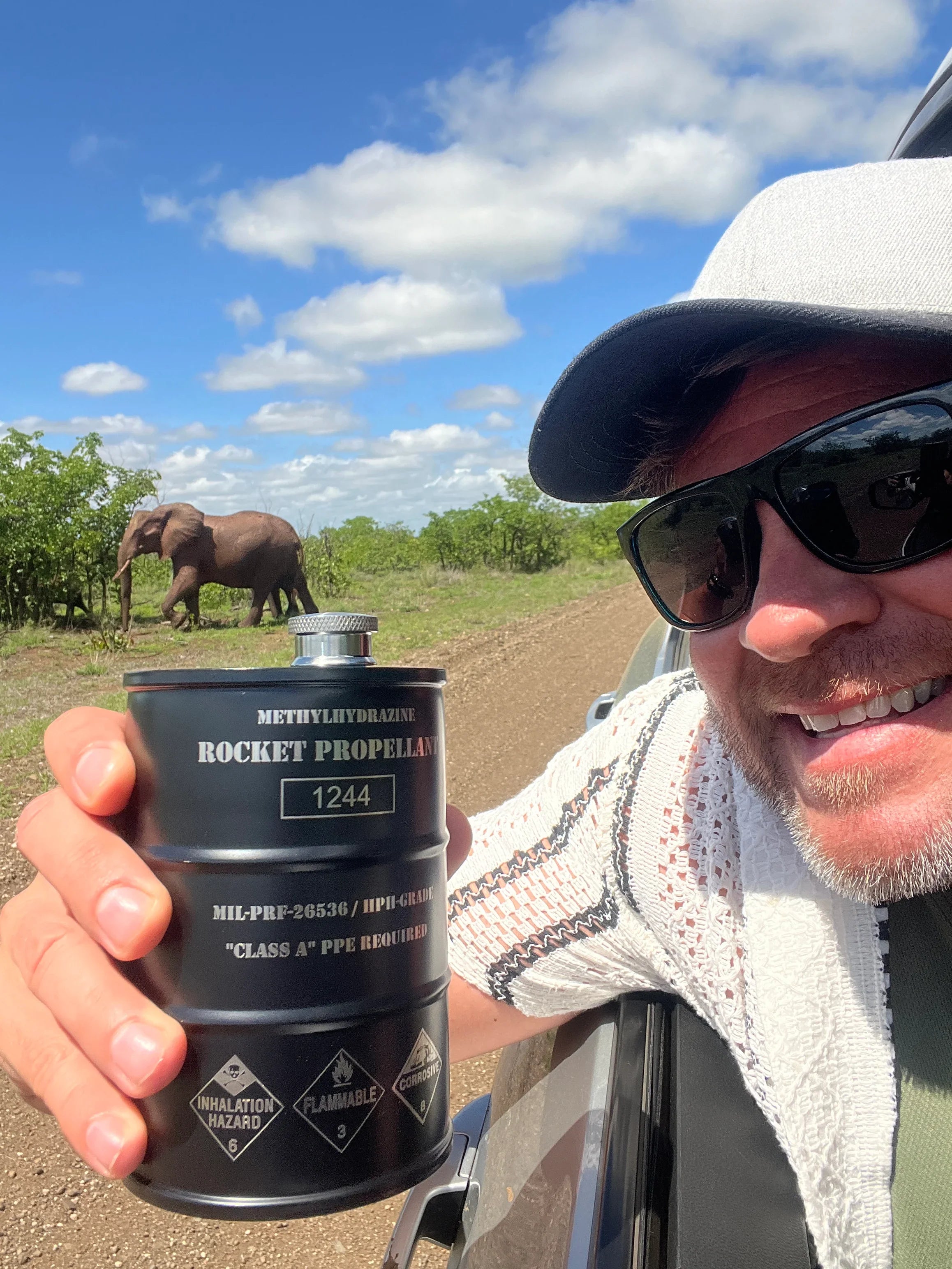 Man wearing sunglasses and cap holding black methylhydrazine rocket propellant canister in safari vehicle with elephant in background