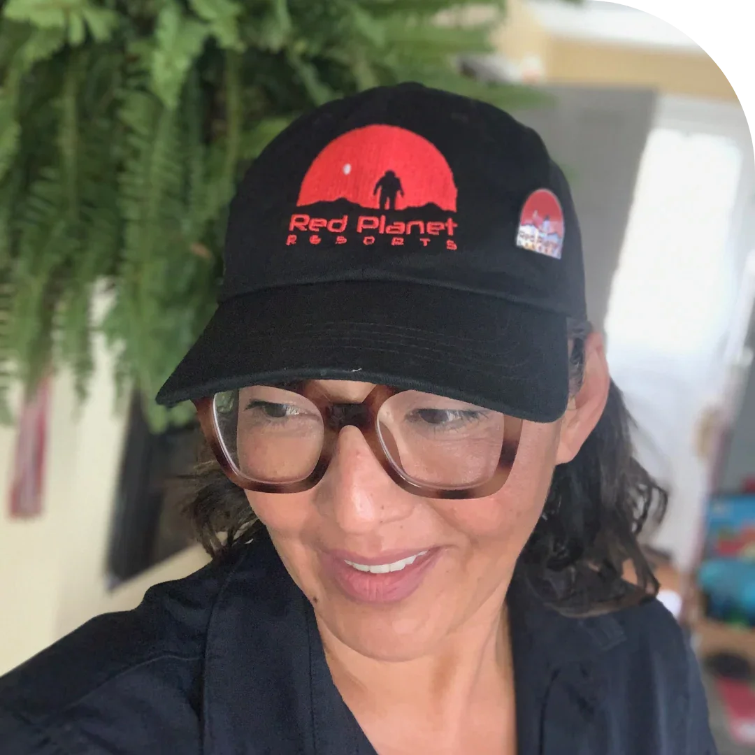 Woman wearing black Red Planet Resorts cap with red logo and large brown glasses indoors