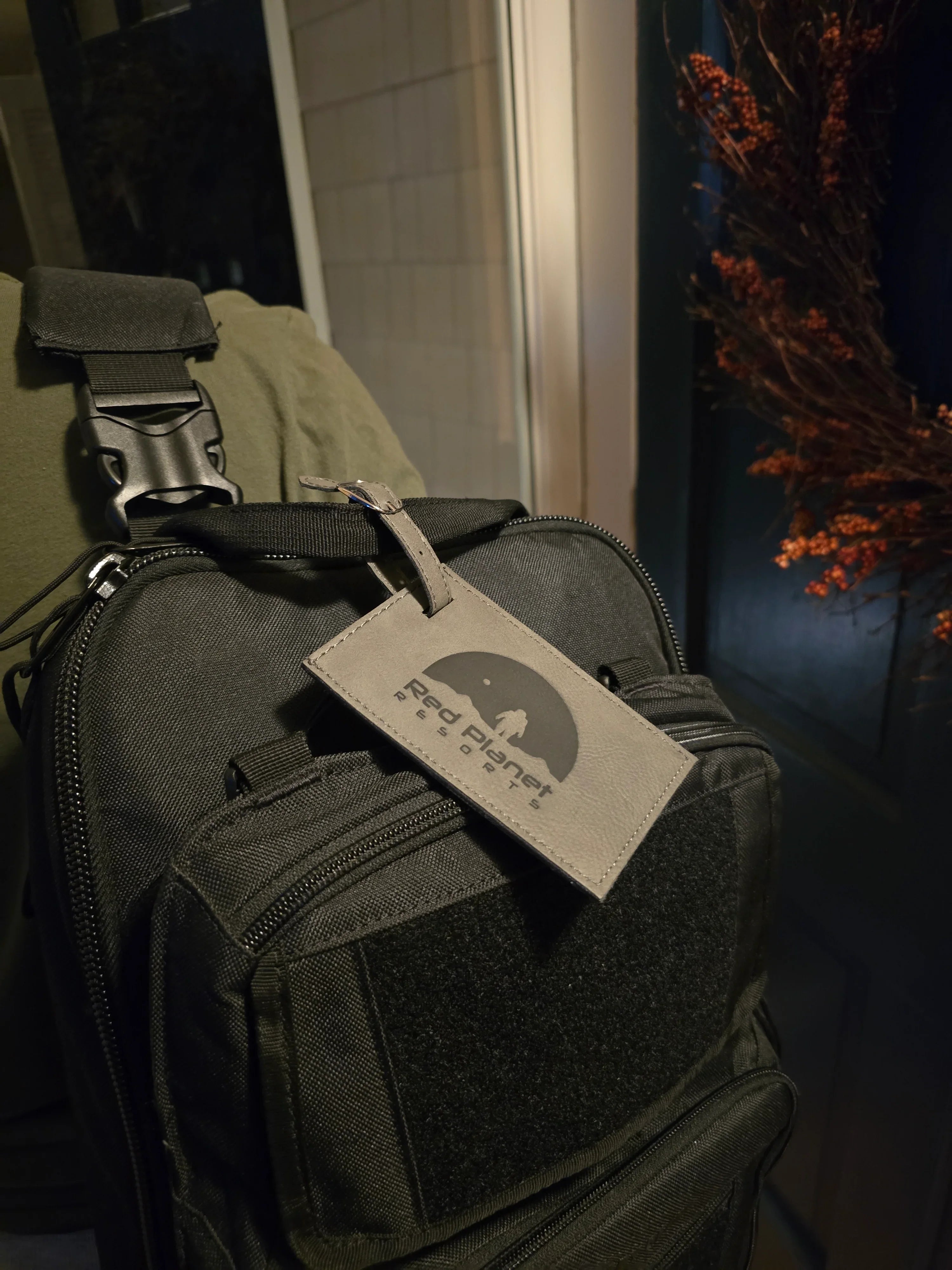 Black tactical backpack with Red Planet Resorts luggage tag hanging, indoor setting with dried floral decor