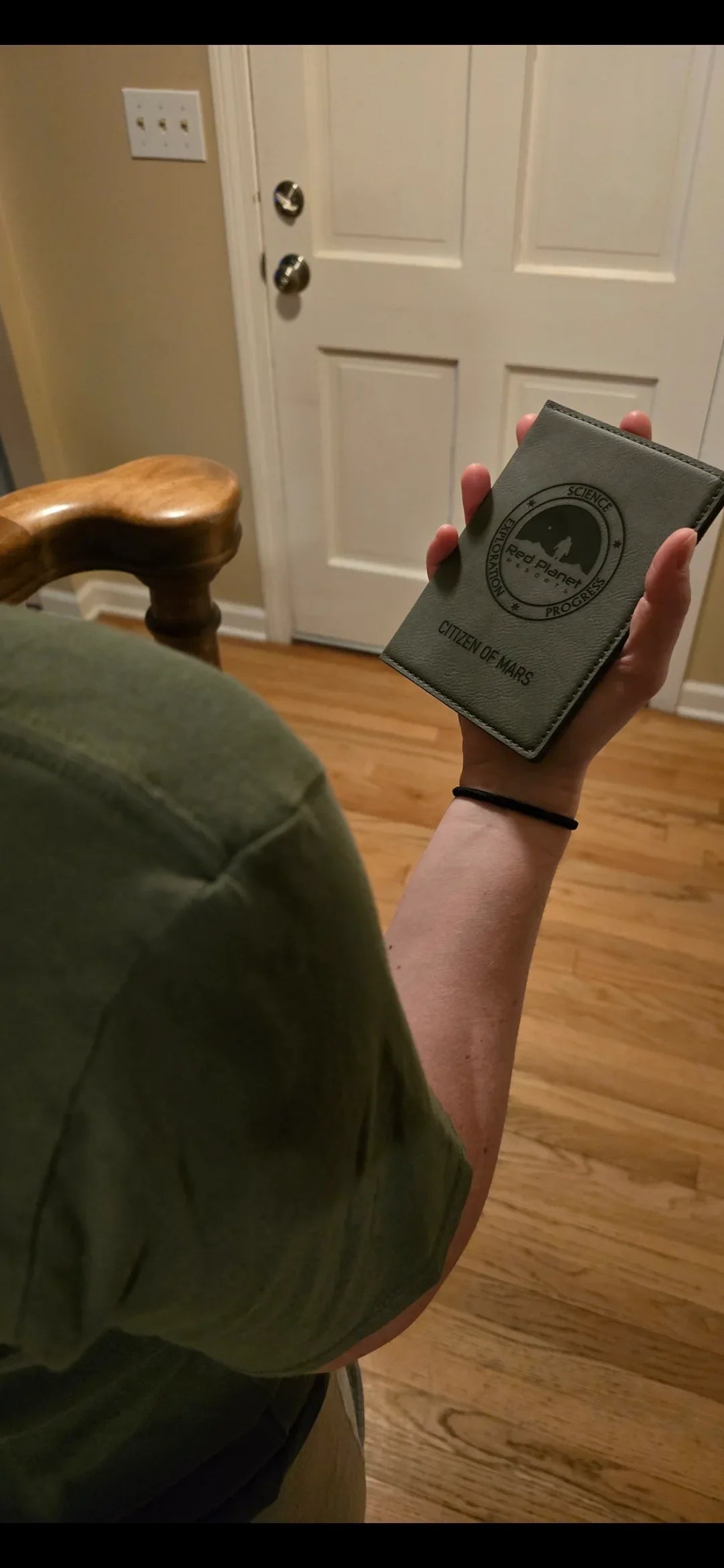 Person holding green booklet labeled Citizen of Mars with Red Planet logo near wooden chair and closed door