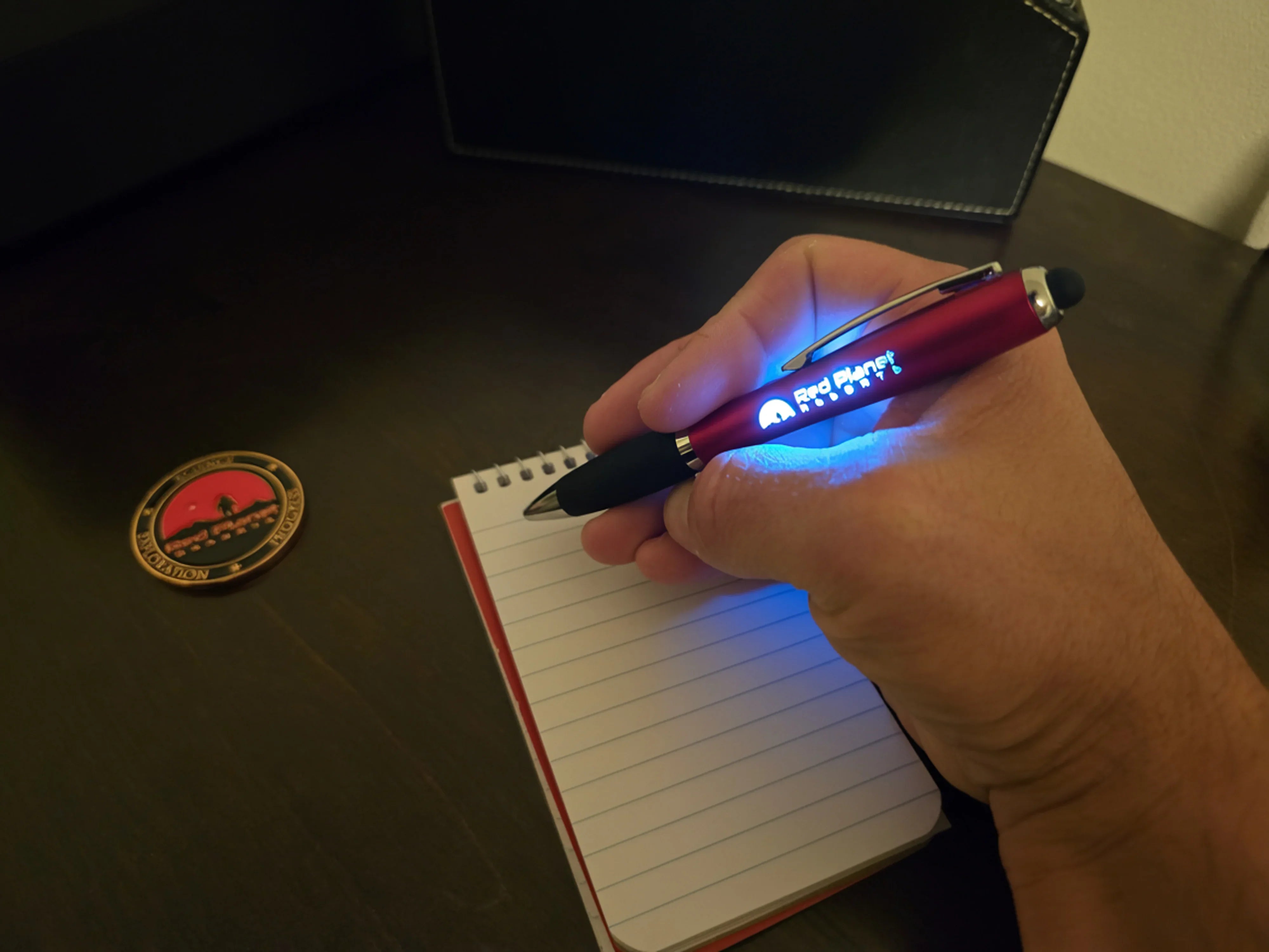 Hand holding a red illuminated Red Planet Robotics pen writing on a small lined notepad on a dark desk
