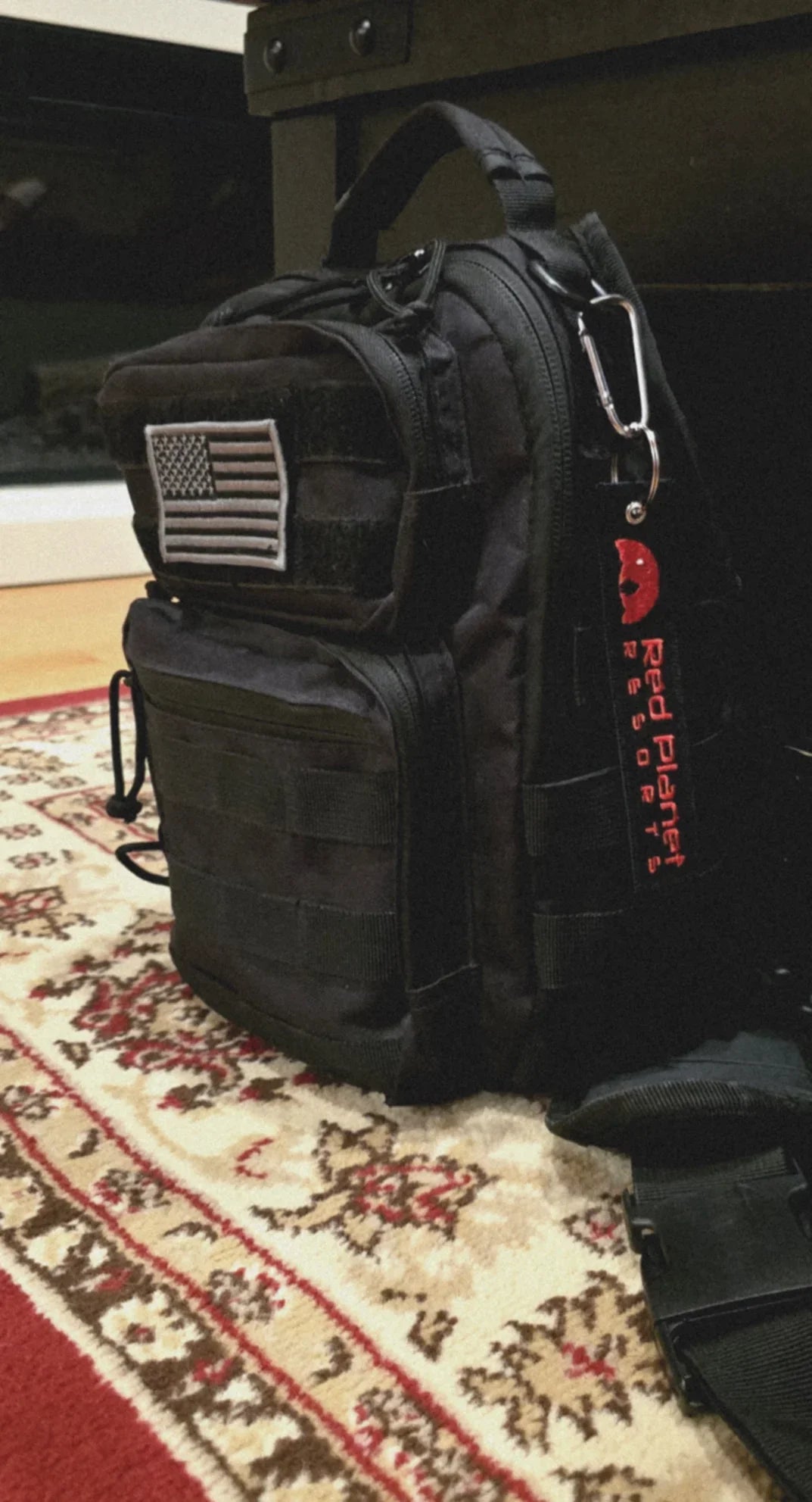 Black tactical backpack with American flag patch and Red Planet Resorts keychain on patterned carpet