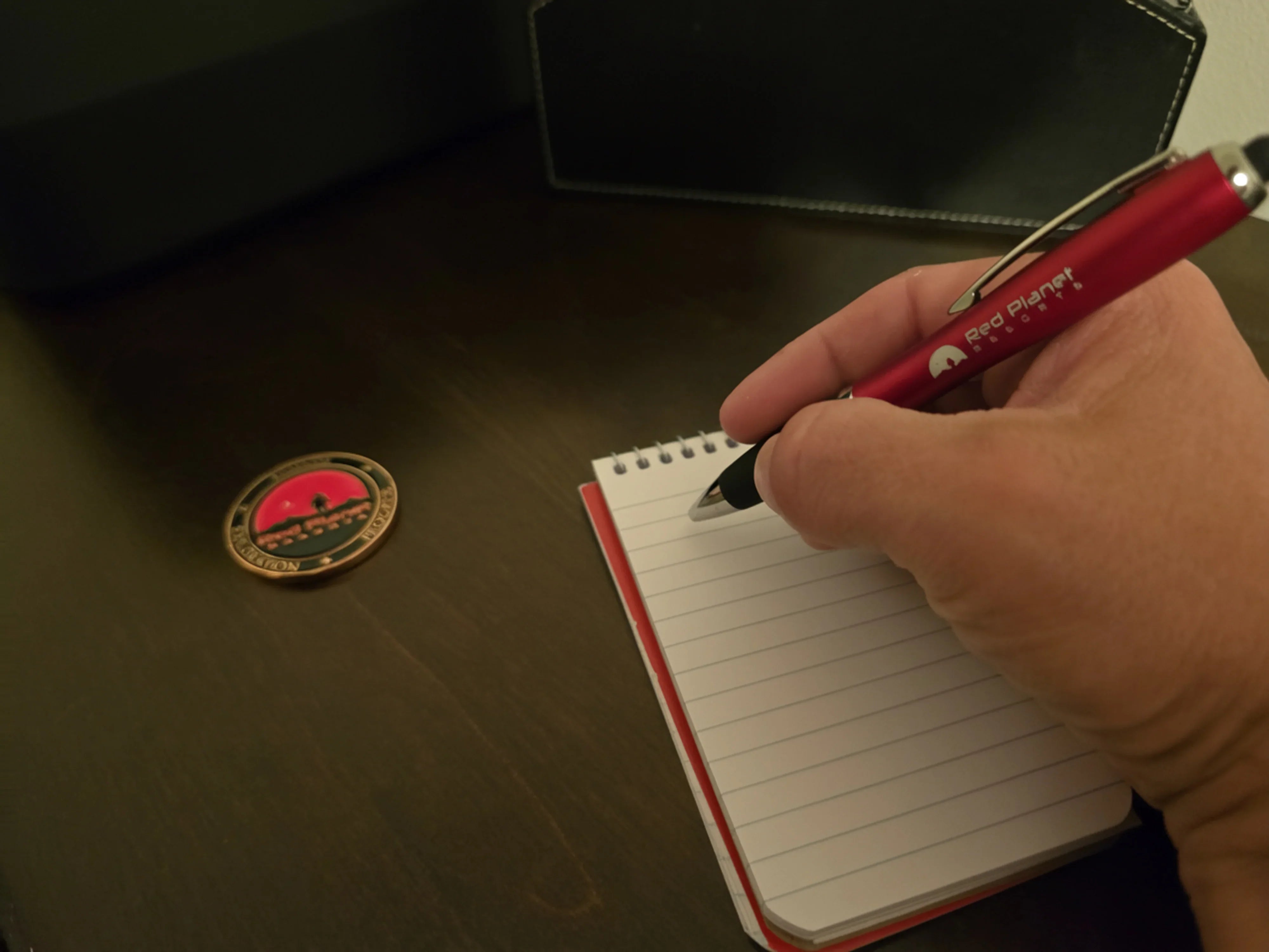 Hand holding a Red Planet Detroit pen poised over a blank spiral notebook on a dark wooden desk
