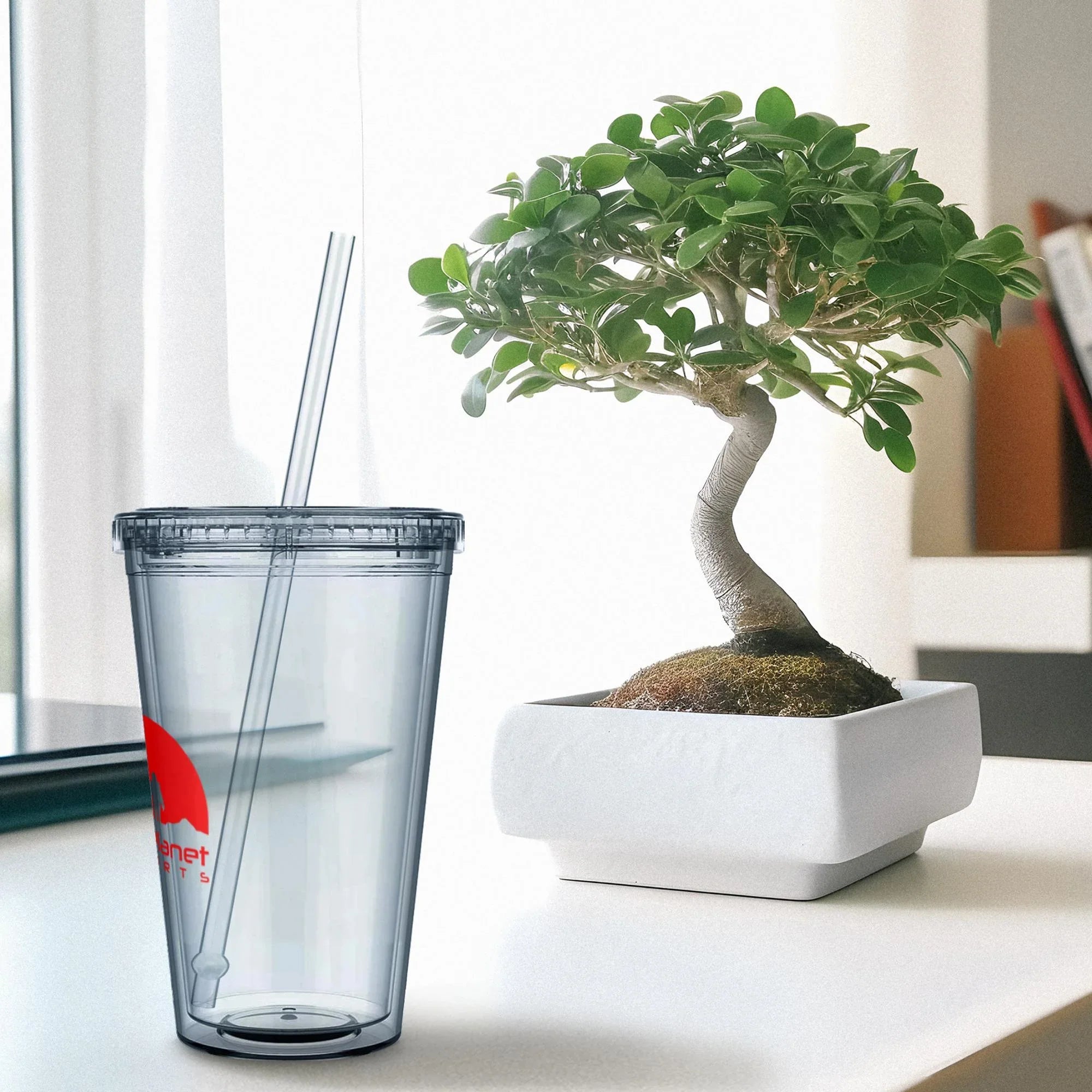 Clear reusable tumbler with lid and straw beside a bonsai tree in a white square pot on a white surface