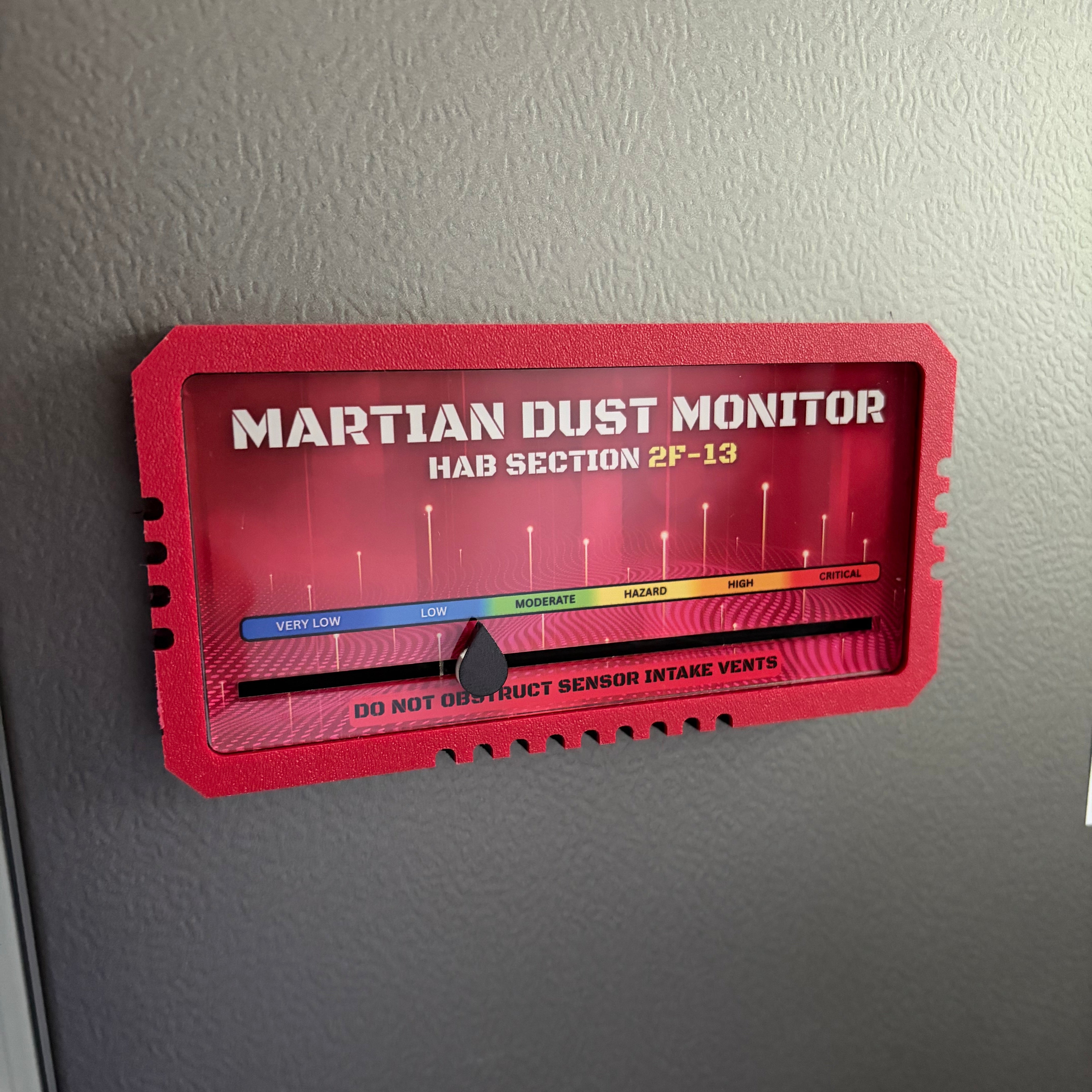 A red 'Martian Dust Monitor' plaque mounted on a dark gray textured wall. The plaque features a red background with a multi-colored horizontal bar (blue, green, yellow, orange) and white text reading 'MARTIAN DUST MONITOR HAB SECTION 2F-13'.
