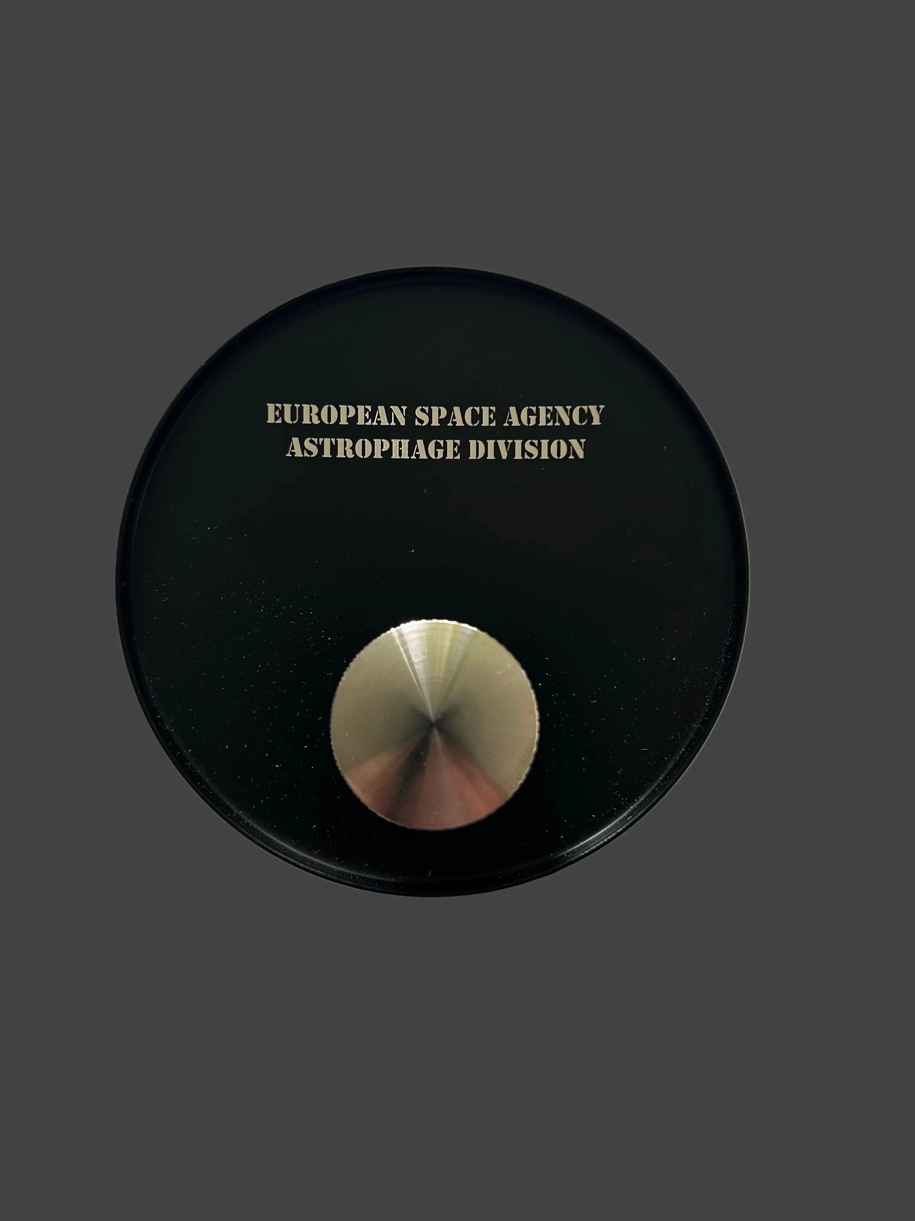 Black circular object with European Space Agency Astrophage Division text and central metallic button