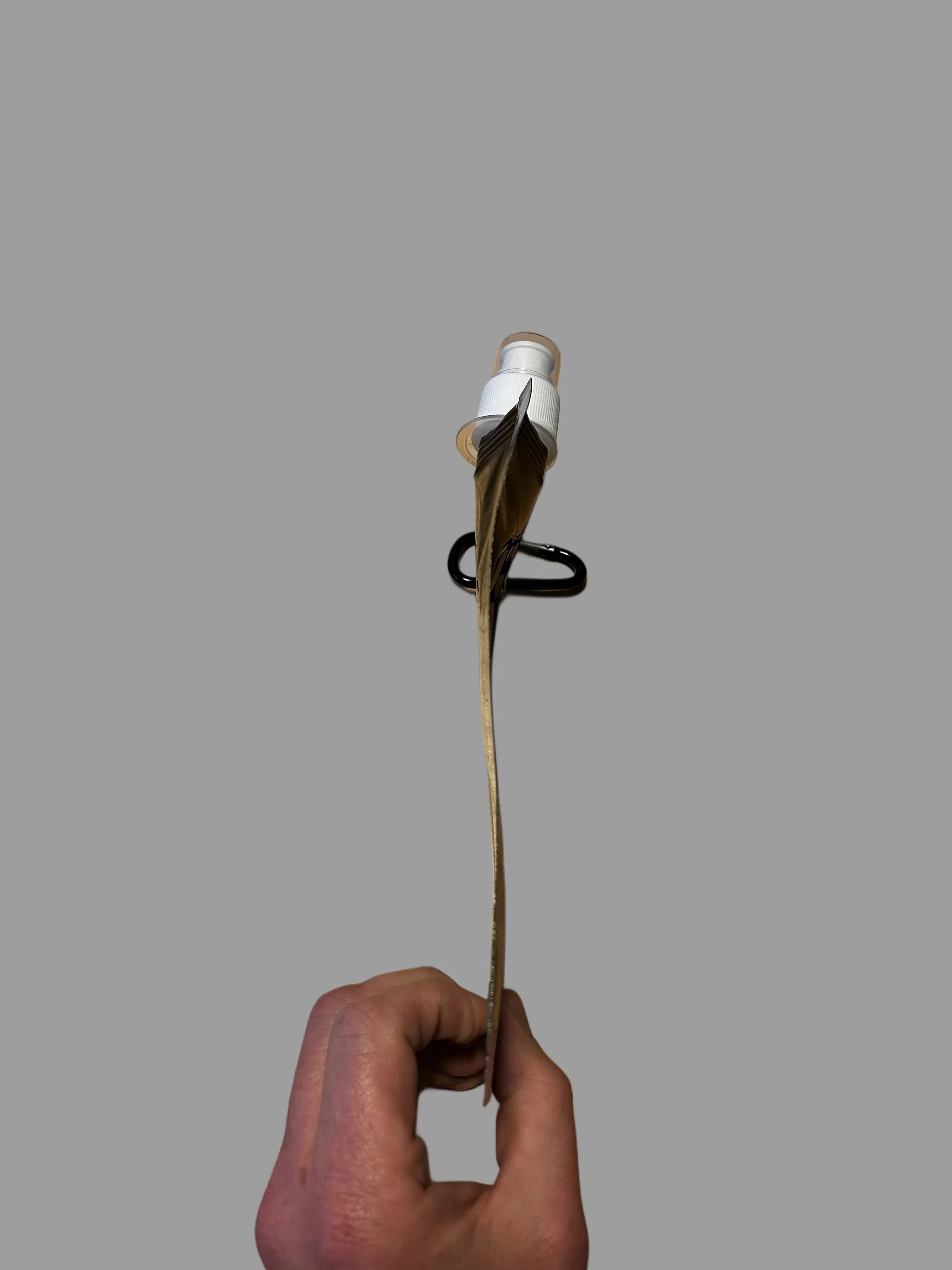 Hand holding a small, thin cardboard package with a white spray nozzle and black carabiner against a gray background