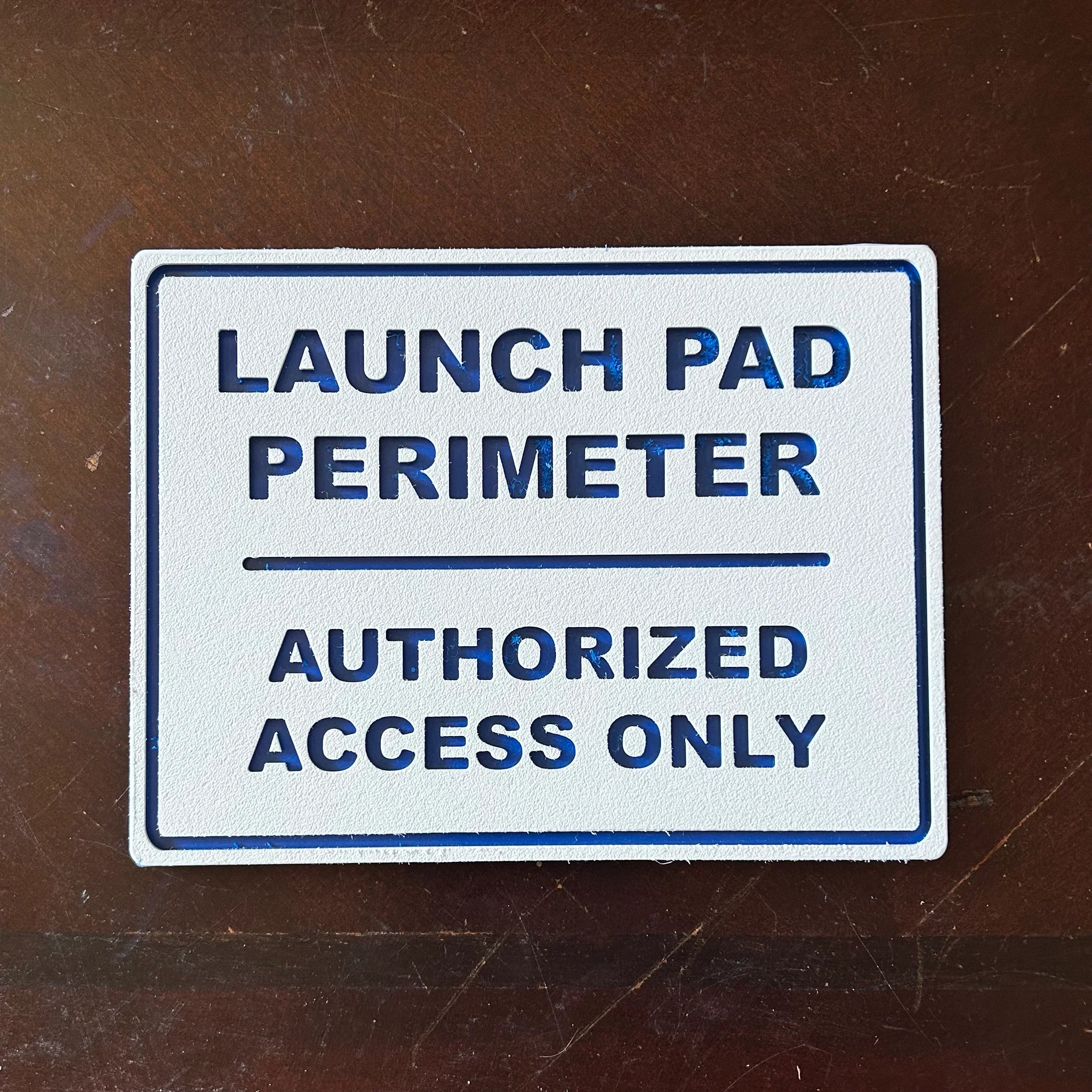 White textured sign with blue text reading Launch Pad Perimeter Authorized Access Only on wooden surface