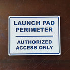 White textured sign with blue text reading Launch Pad Perimeter Authorized Access Only on wooden surface