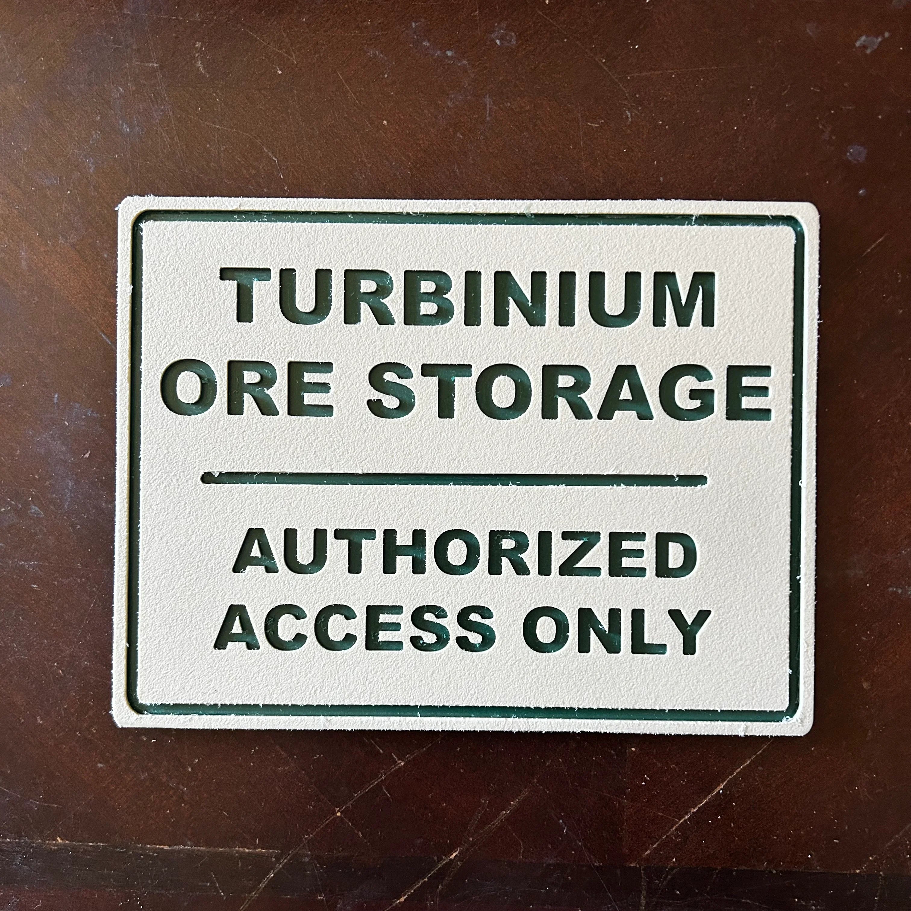 Beige sign with green text reading Turbinum Ore Storage Authorized Access Only on brown surface