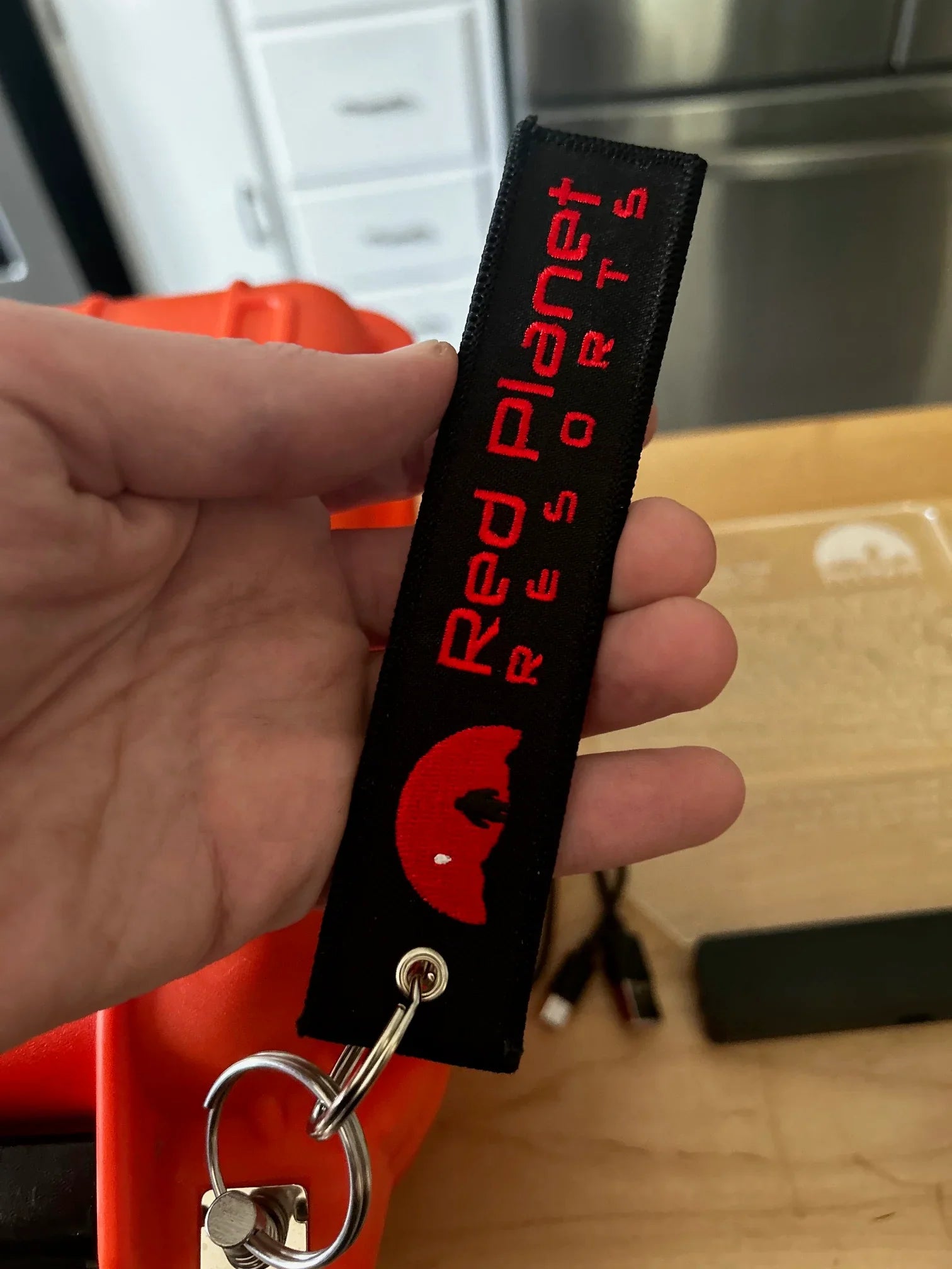 Hand holding a Red Planet Resorts keychain with red logo and text on black fabric background