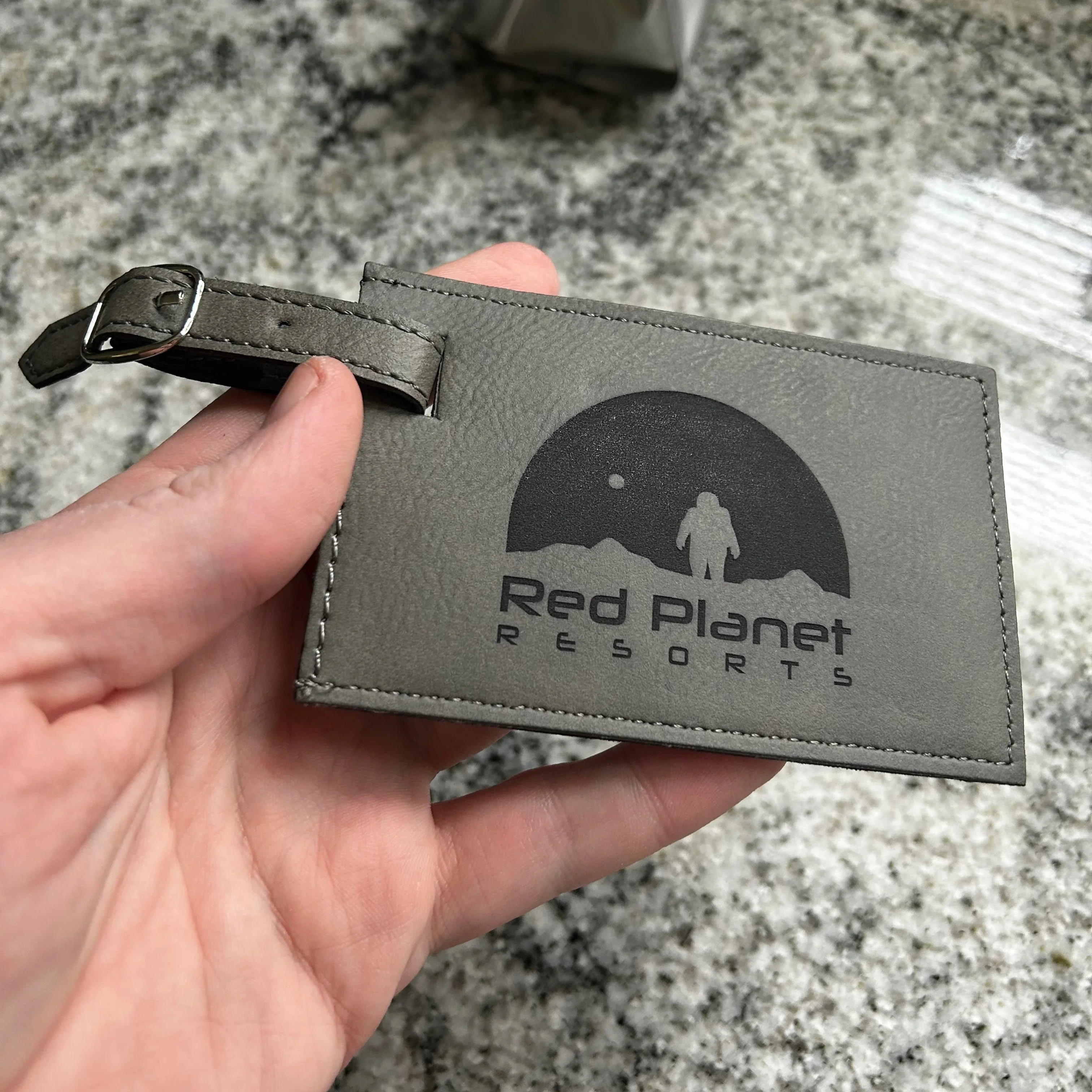 Hand holding gray leather luggage tag with Red Planet Resorts logo on granite surface