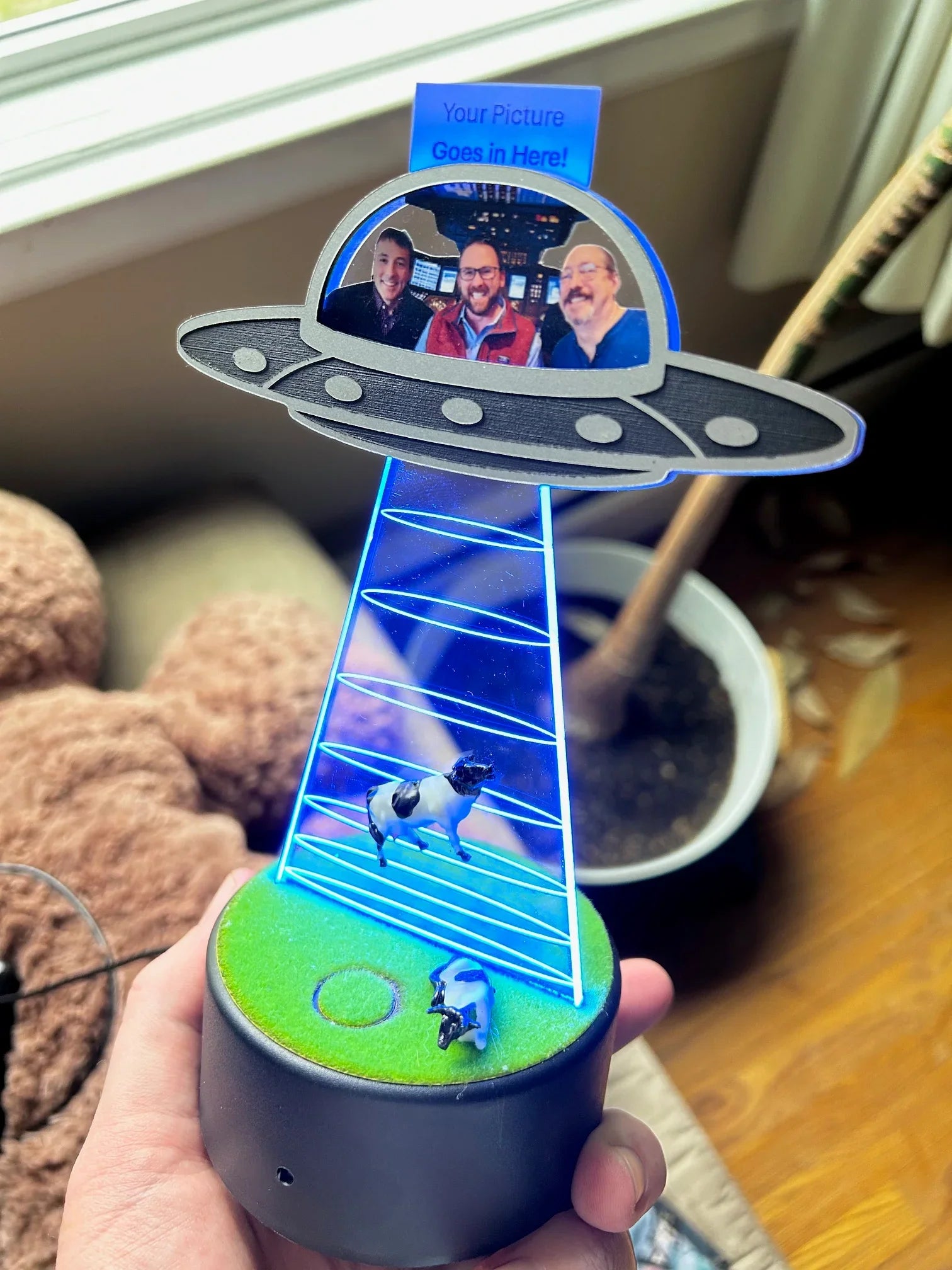 Hand holding UFO-themed LED lamp with abducted cow and photo insert of three men in spaceship cockpit