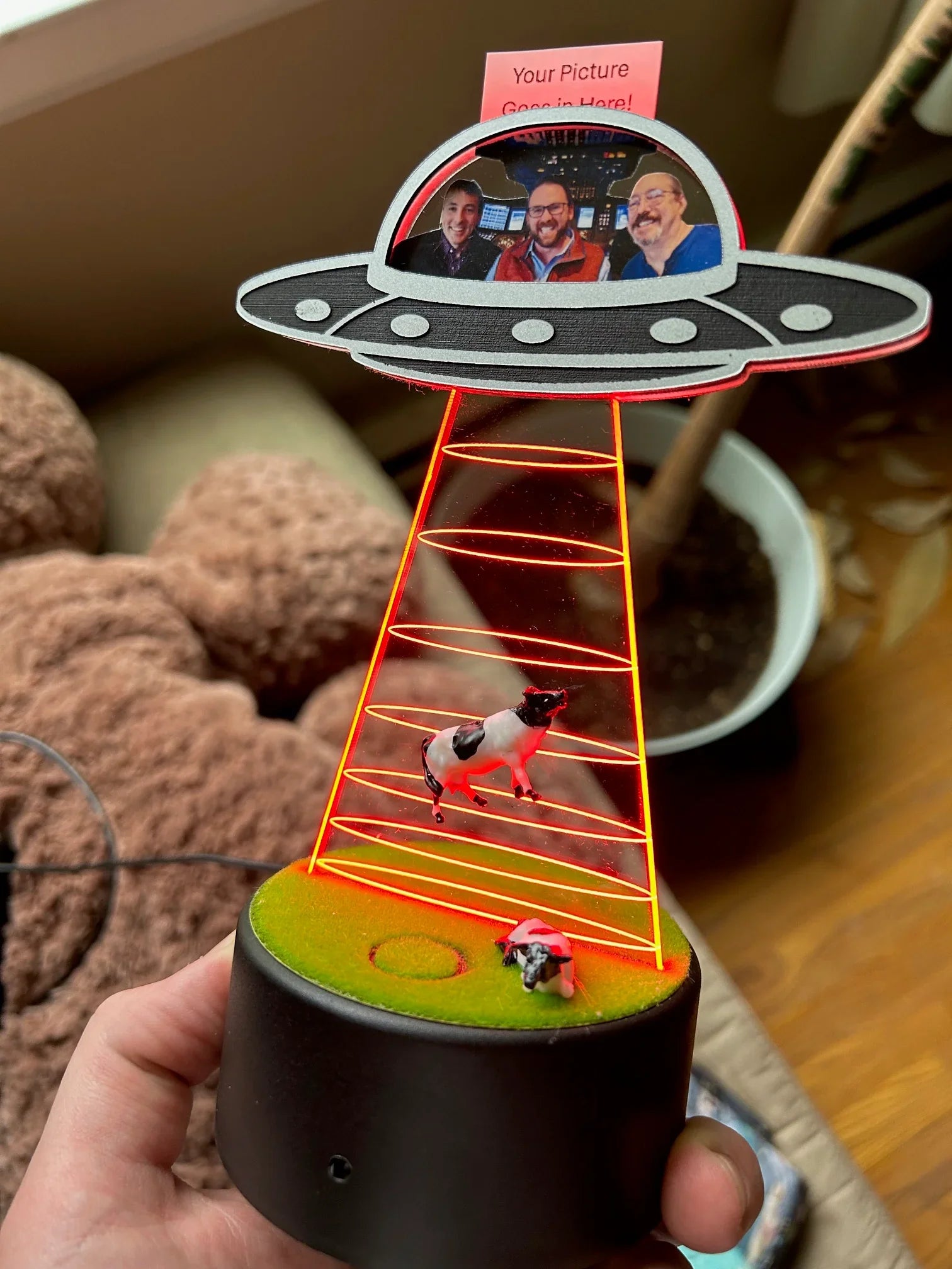 Hand holding a UFO-themed LED lamp with holographic cow abduction and photo of three men in spaceship cockpit