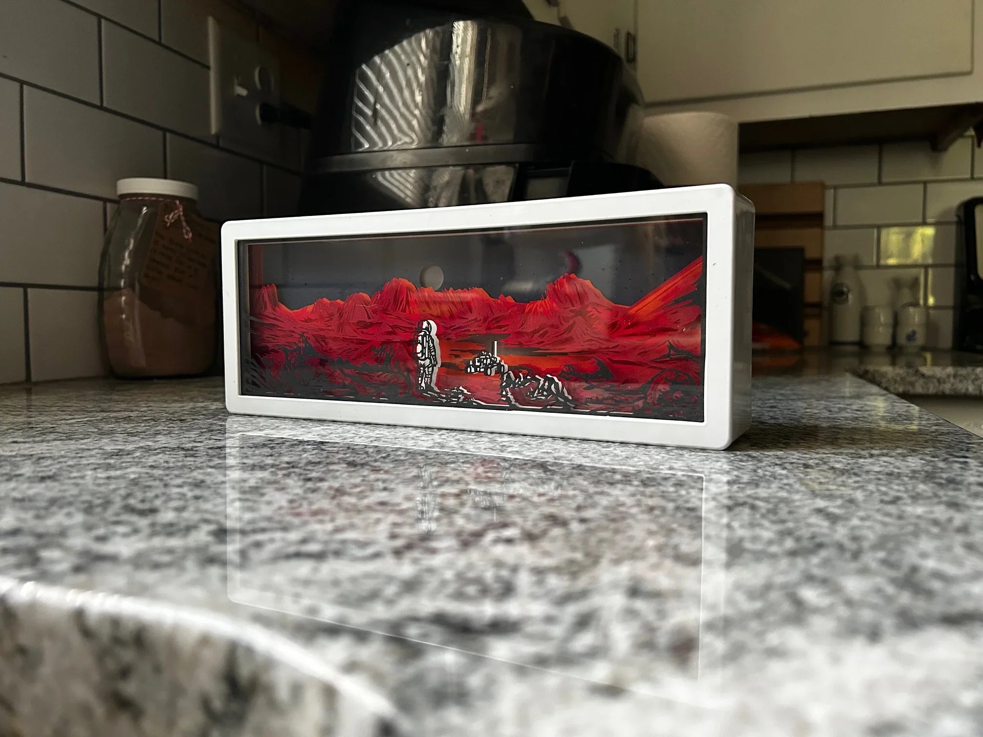 Rectangular white shadow box on granite counter showing an astronaut standing on a red Martian landscape with mountains and a rover