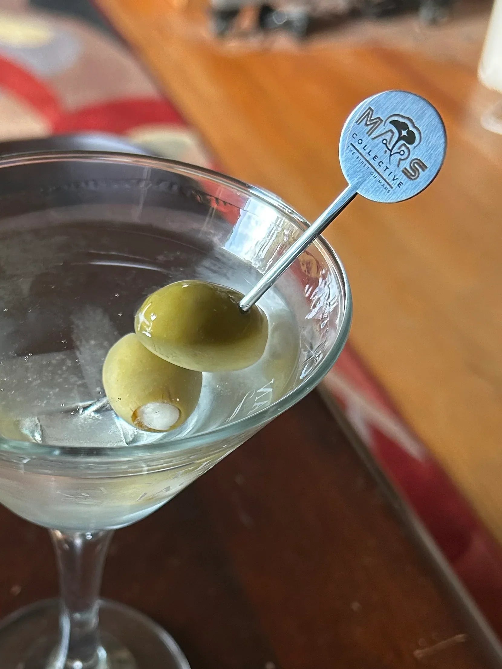 Martini glass with two green olives on a branded Mars Collective cocktail pick on a wooden table