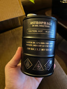 Hand holding a black metal container labeled Astrophage in oil solution with caution hot 96.415°C and hazard symbols