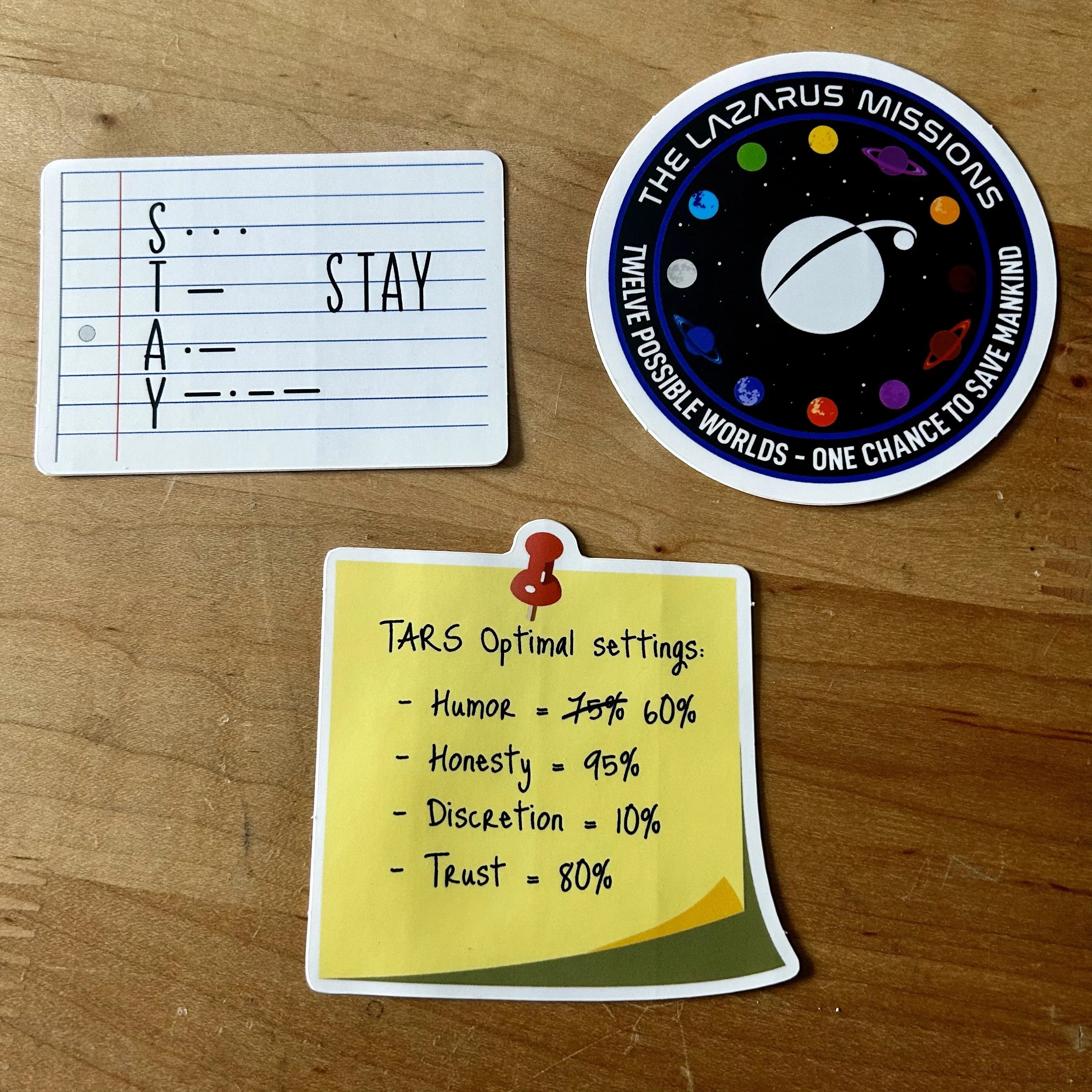 Three Interstellar-themed stickers on wood: Morse code for STAY, Lazarus Missions space patch, and TARS settings note