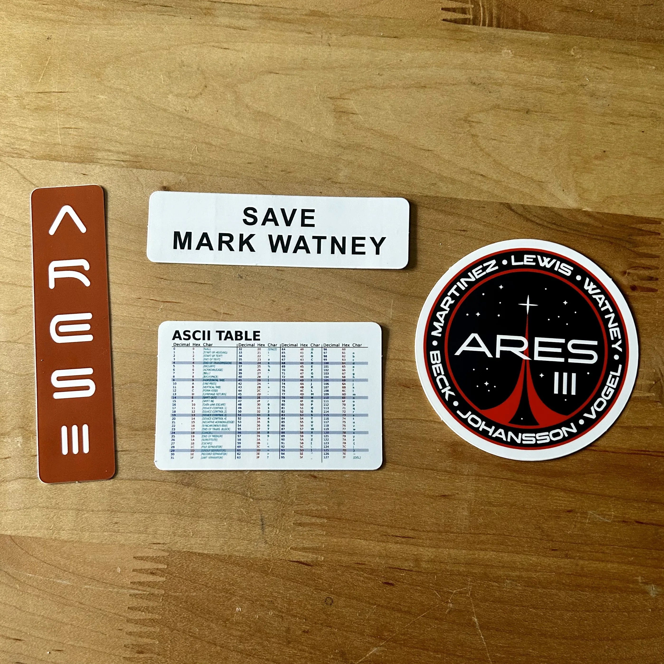 Mars mission themed stickers including Ares III logo, Save Mark Watney, Mars label, and ASCII table on wooden surface