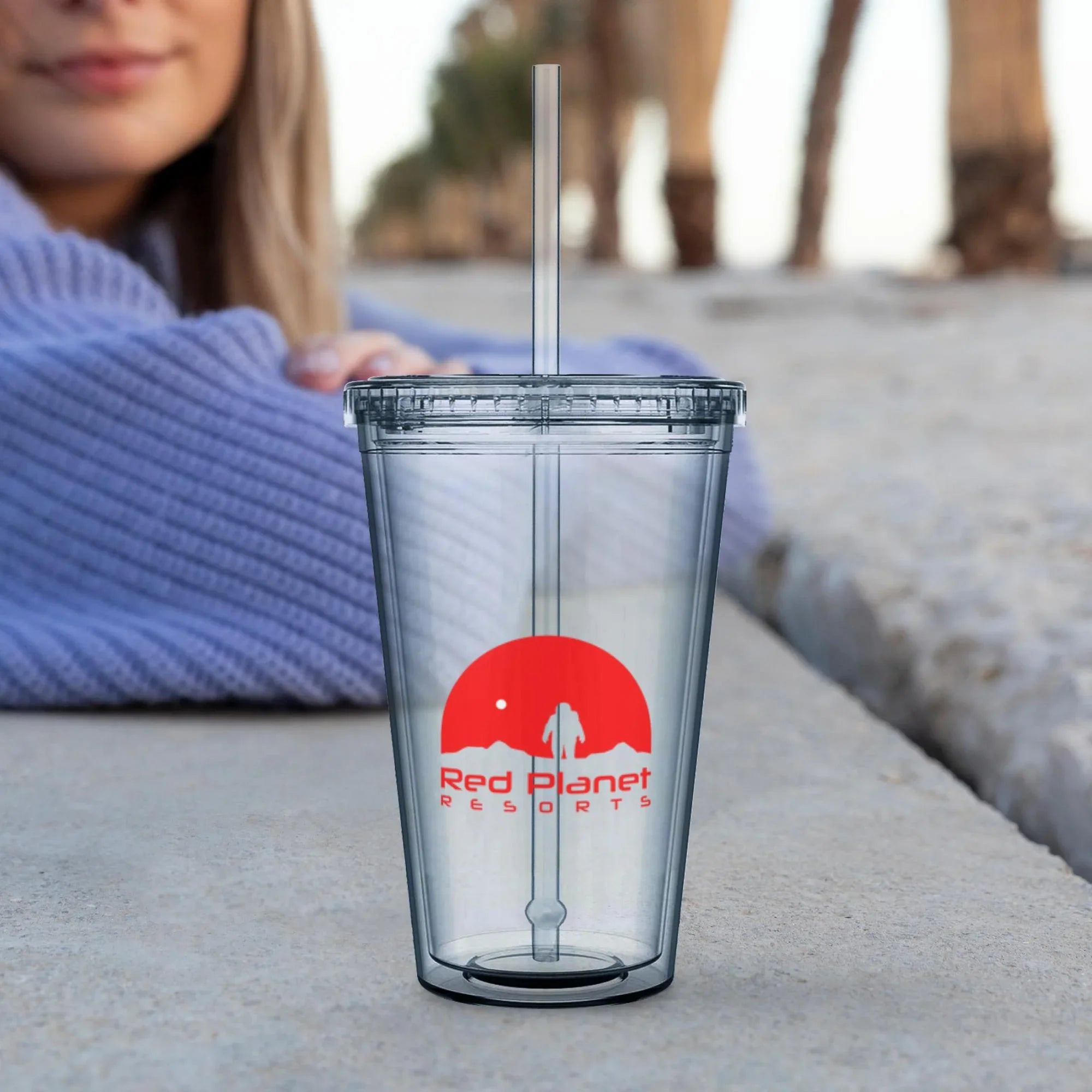Clear reusable plastic tumbler with Red Planet Resorts logo and straw on outdoor surface