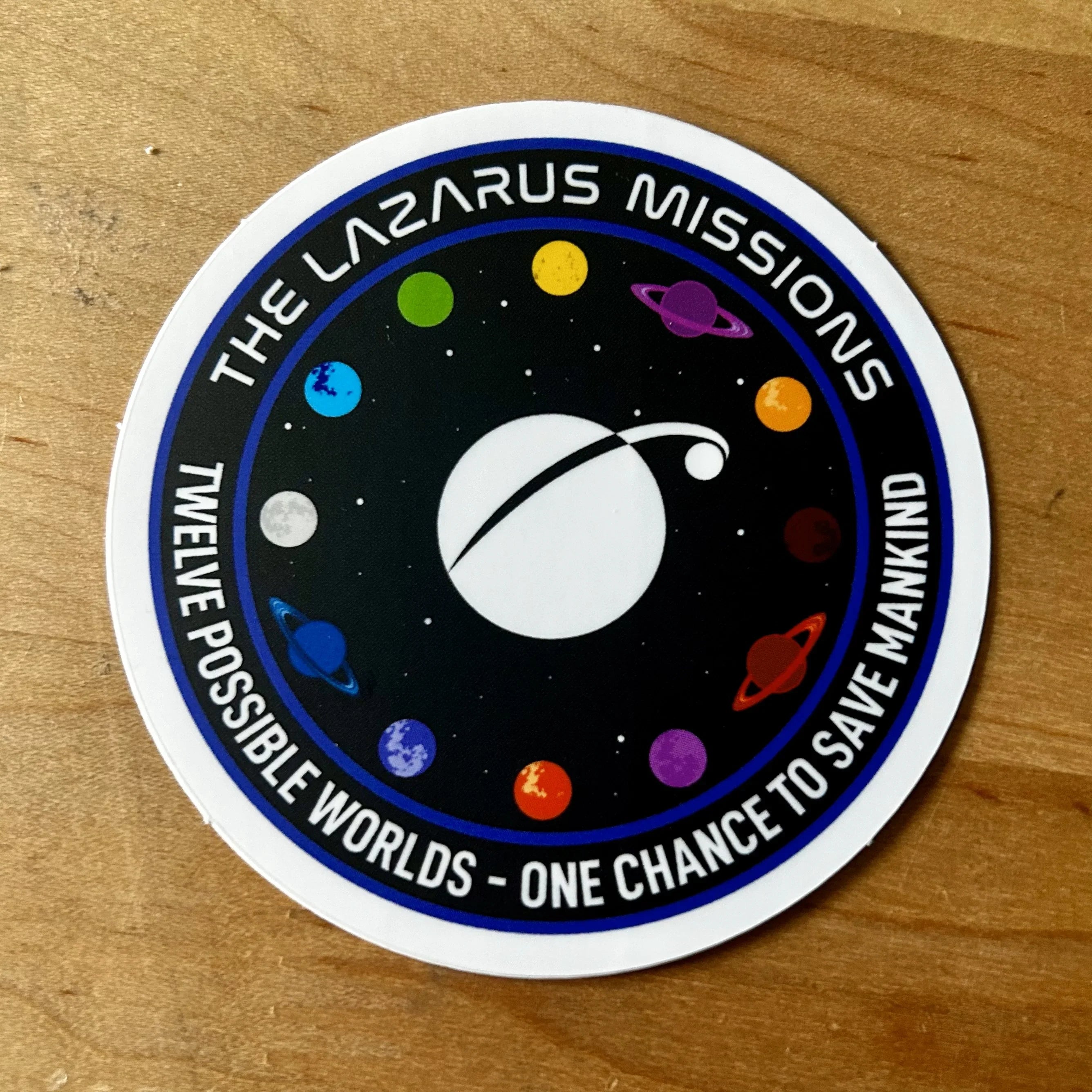 The Lazarus Missions circular space mission patch with colorful planets and the motto on a wooden surface