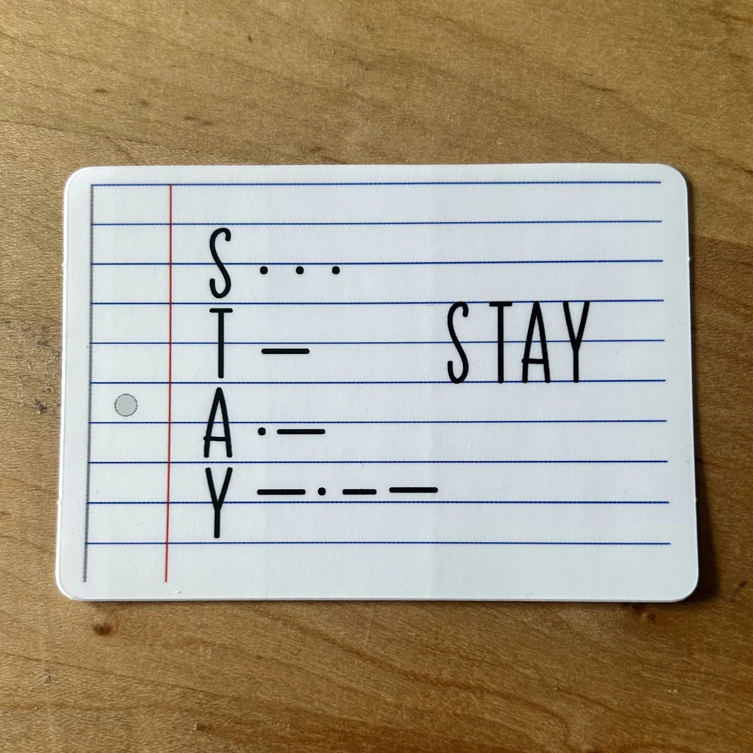 Flashcard with the word STAY spelled out with letters and Morse code on a lined white card on a wooden surface