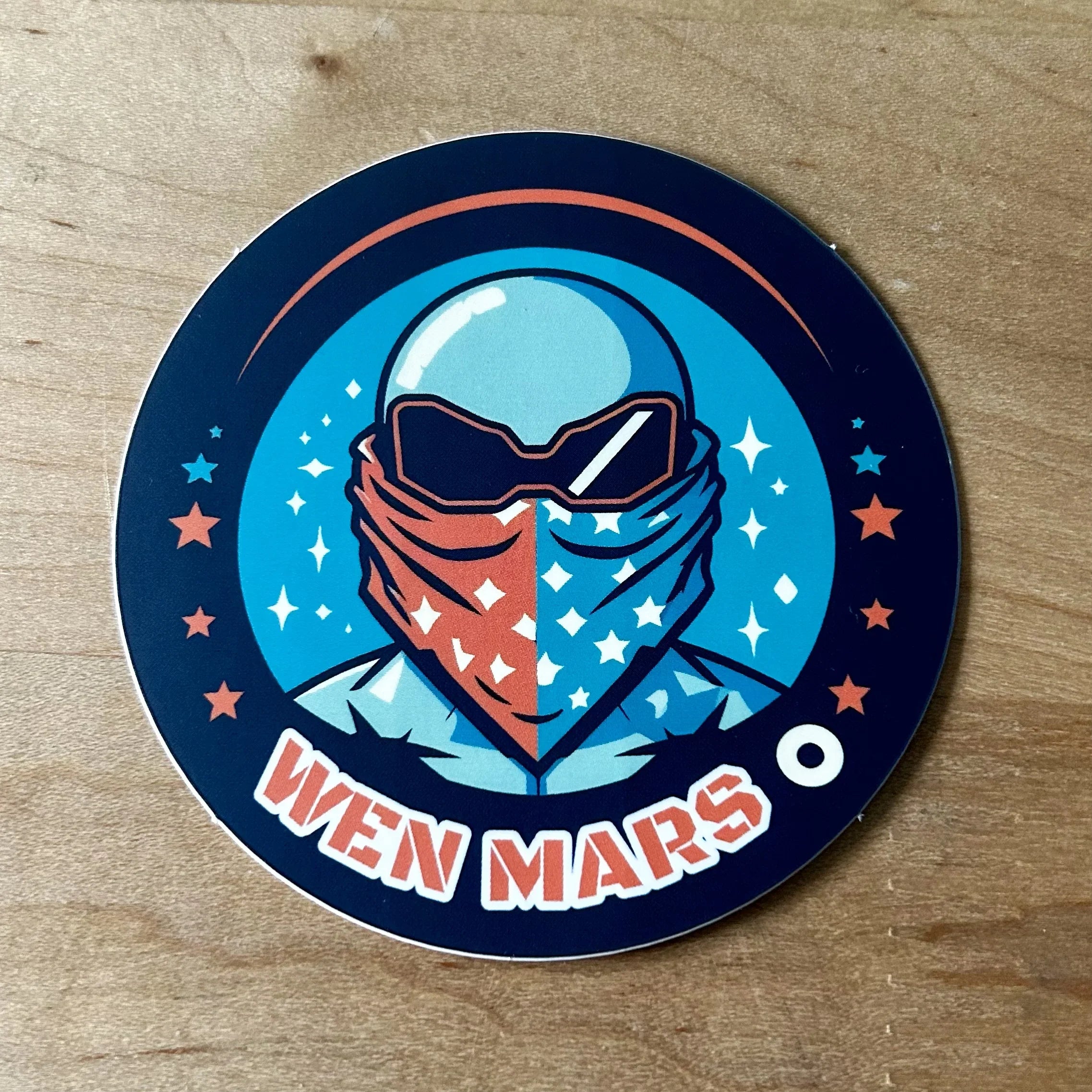 Circular sticker with astronaut wearing split red and blue star-patterned bandana and reflective visor, text "WEN MARS"