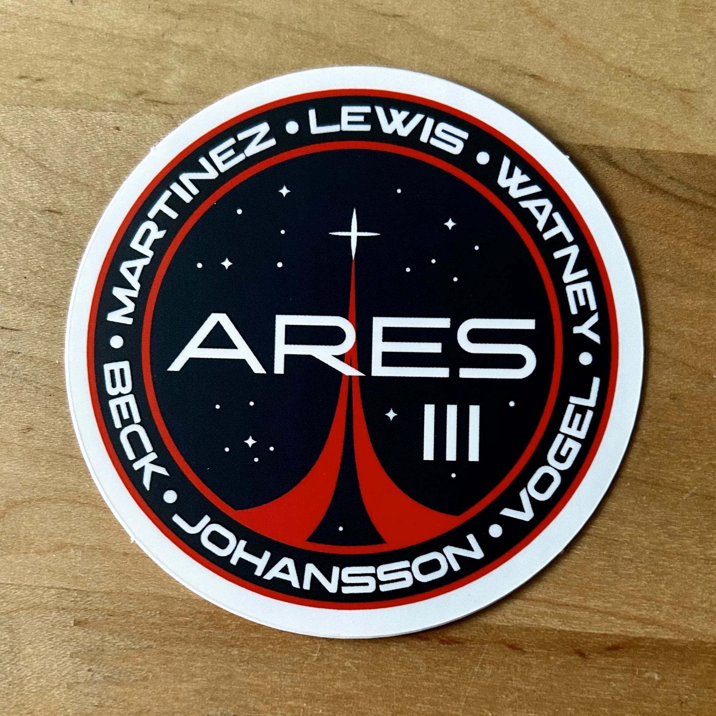 Round Ares III mission patch sticker with stars and red stylized rocket on wood surface