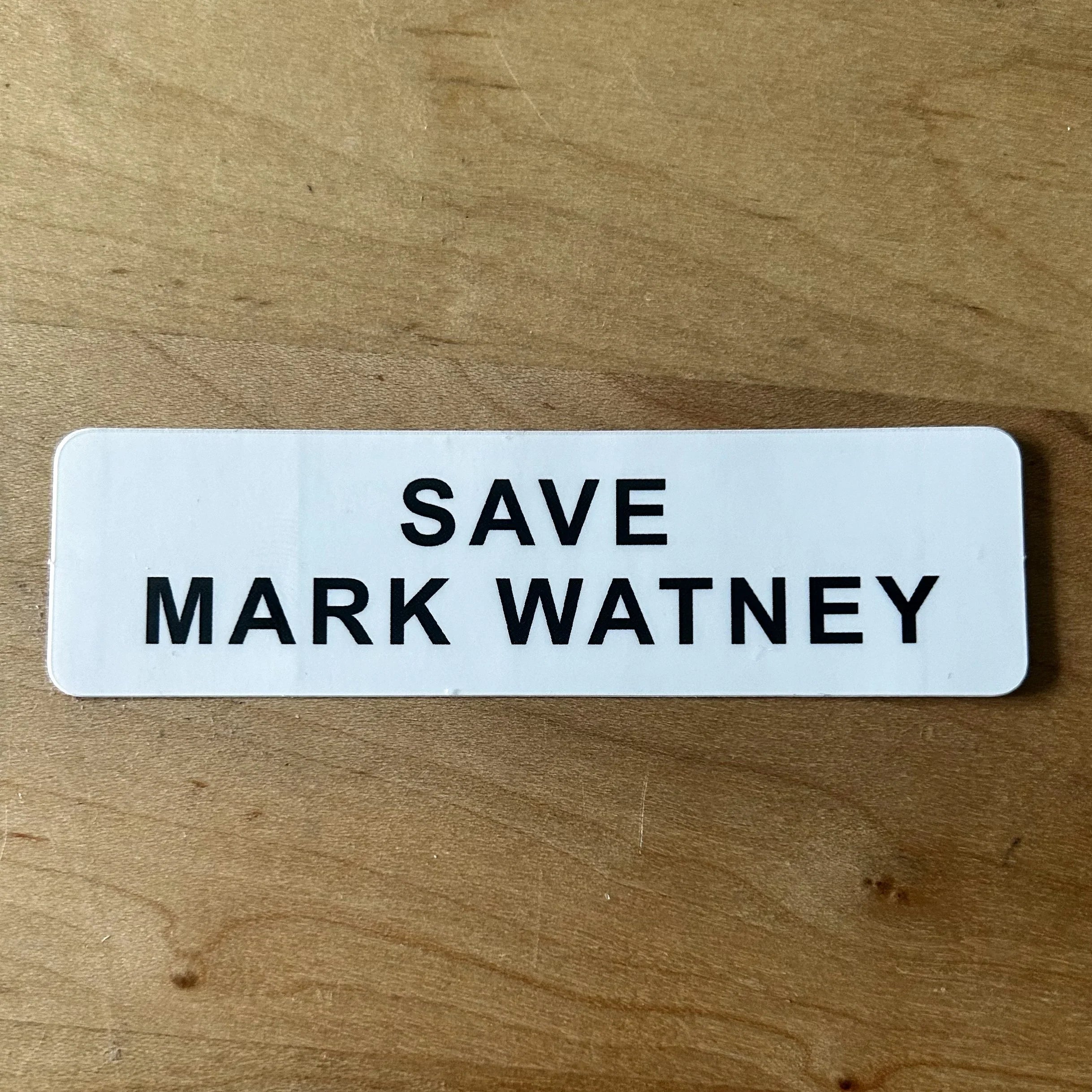 White rectangular sticker with bold black text saying Save Mark Watney on wooden surface