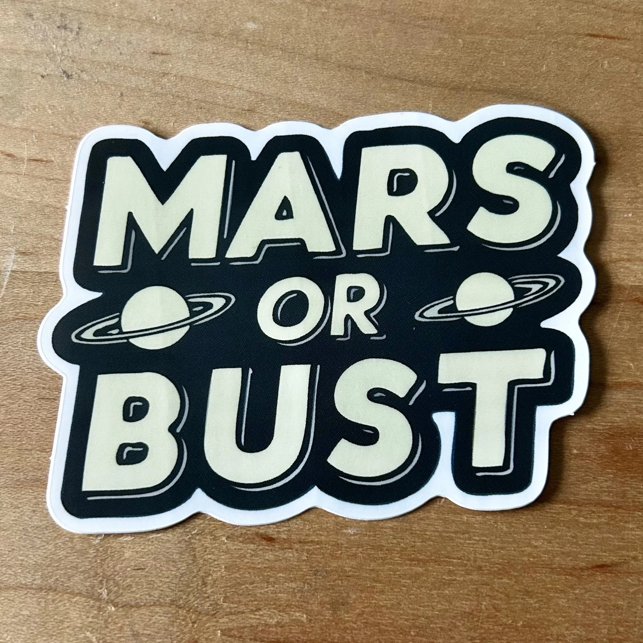 Mars or Bust sticker with planets and rings on wooden surface