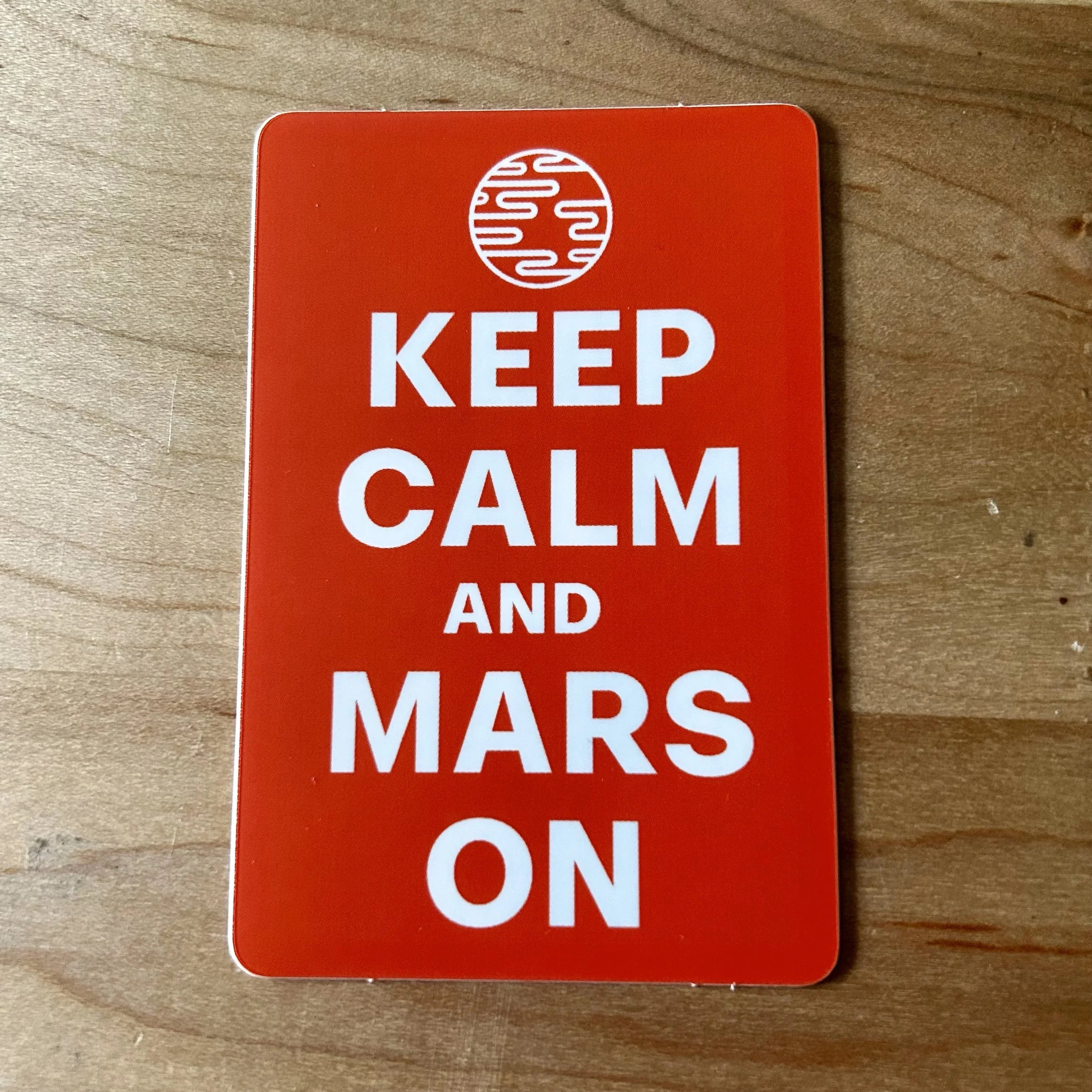 Red card on wooden surface with white text Keep Calm and Mars On and planet Mars icon