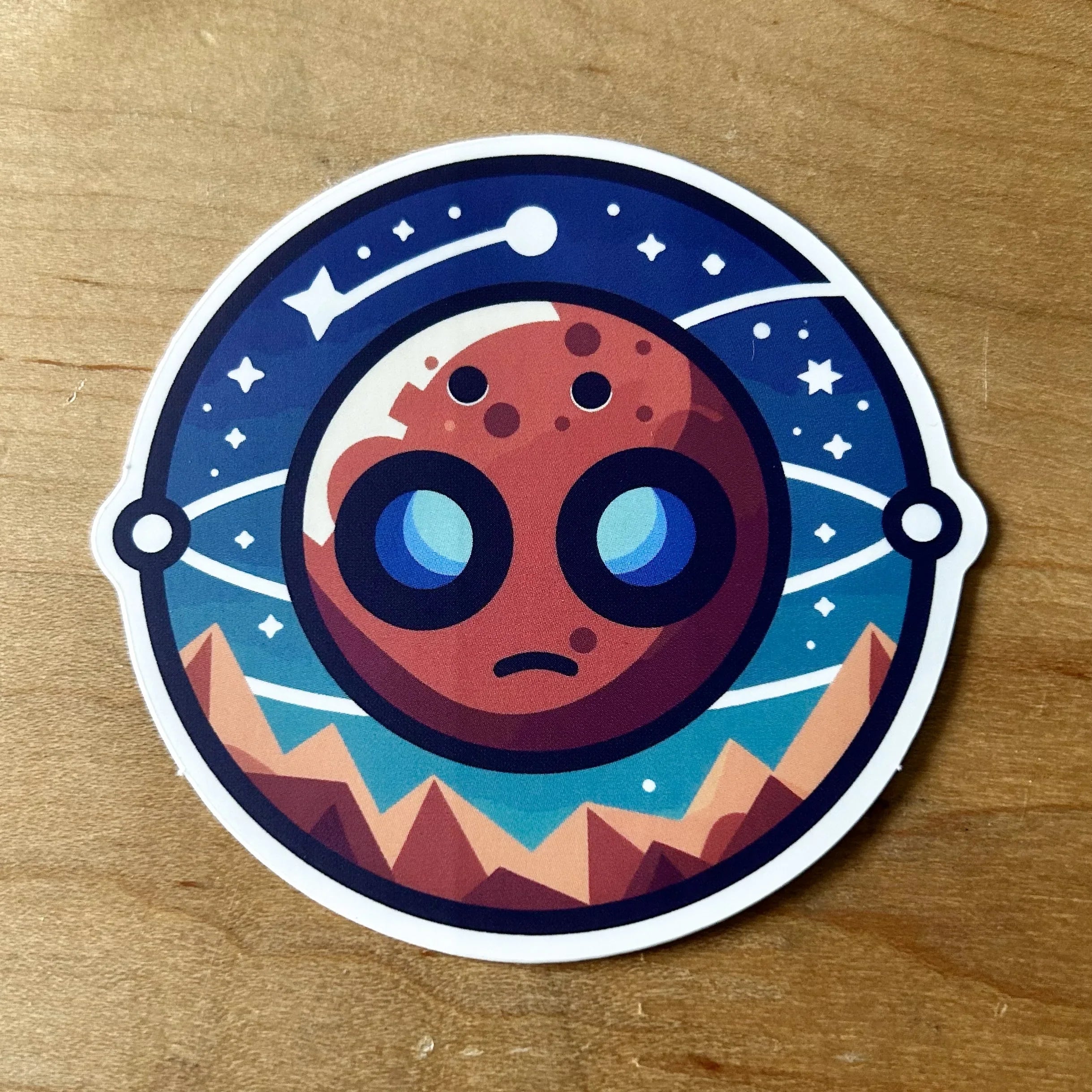 Round sticker of a red planet with large eyes, stars, orbit lines, and mountain landscape on wood background