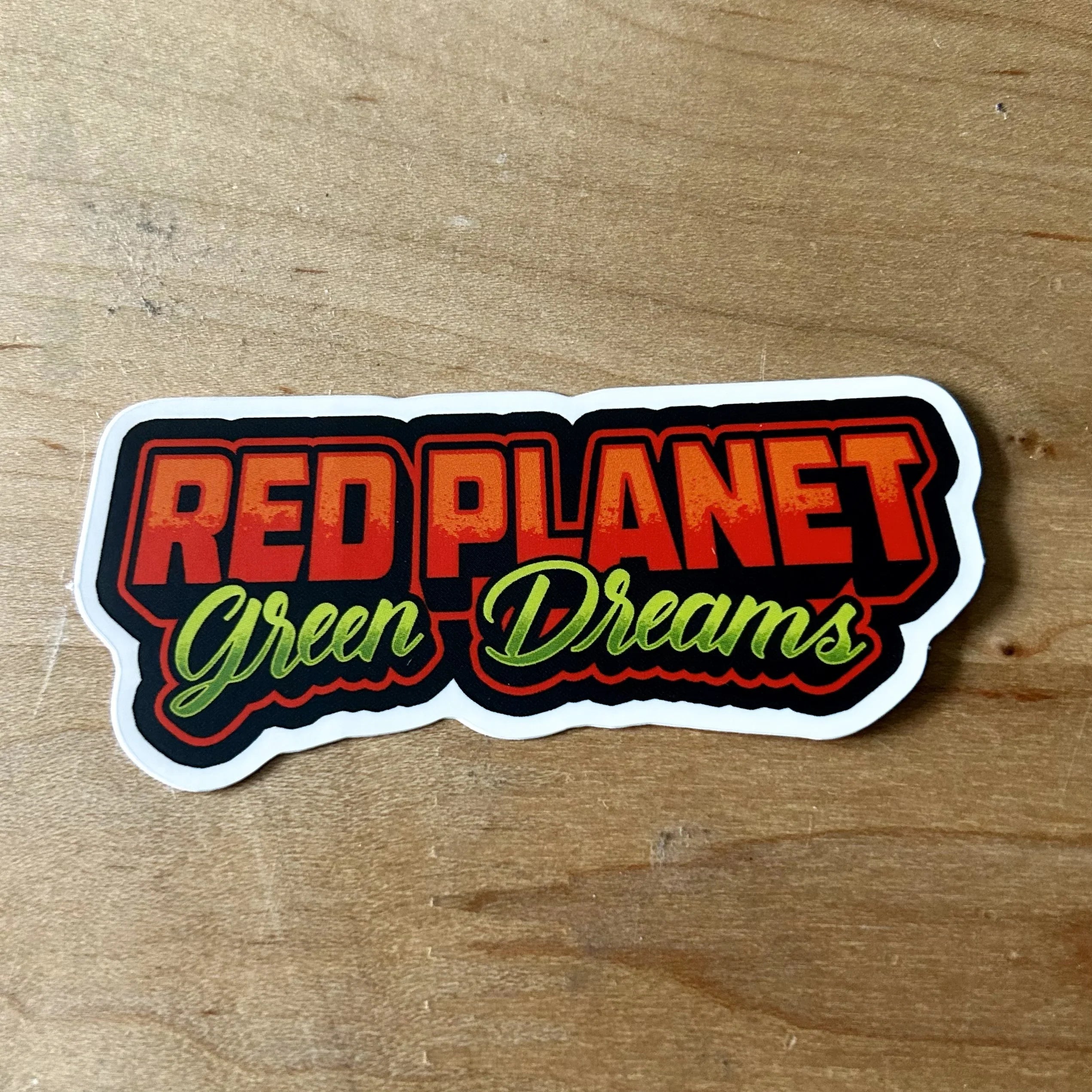 Red Planet Green Dreams sticker with bold red and green text on wooden surface