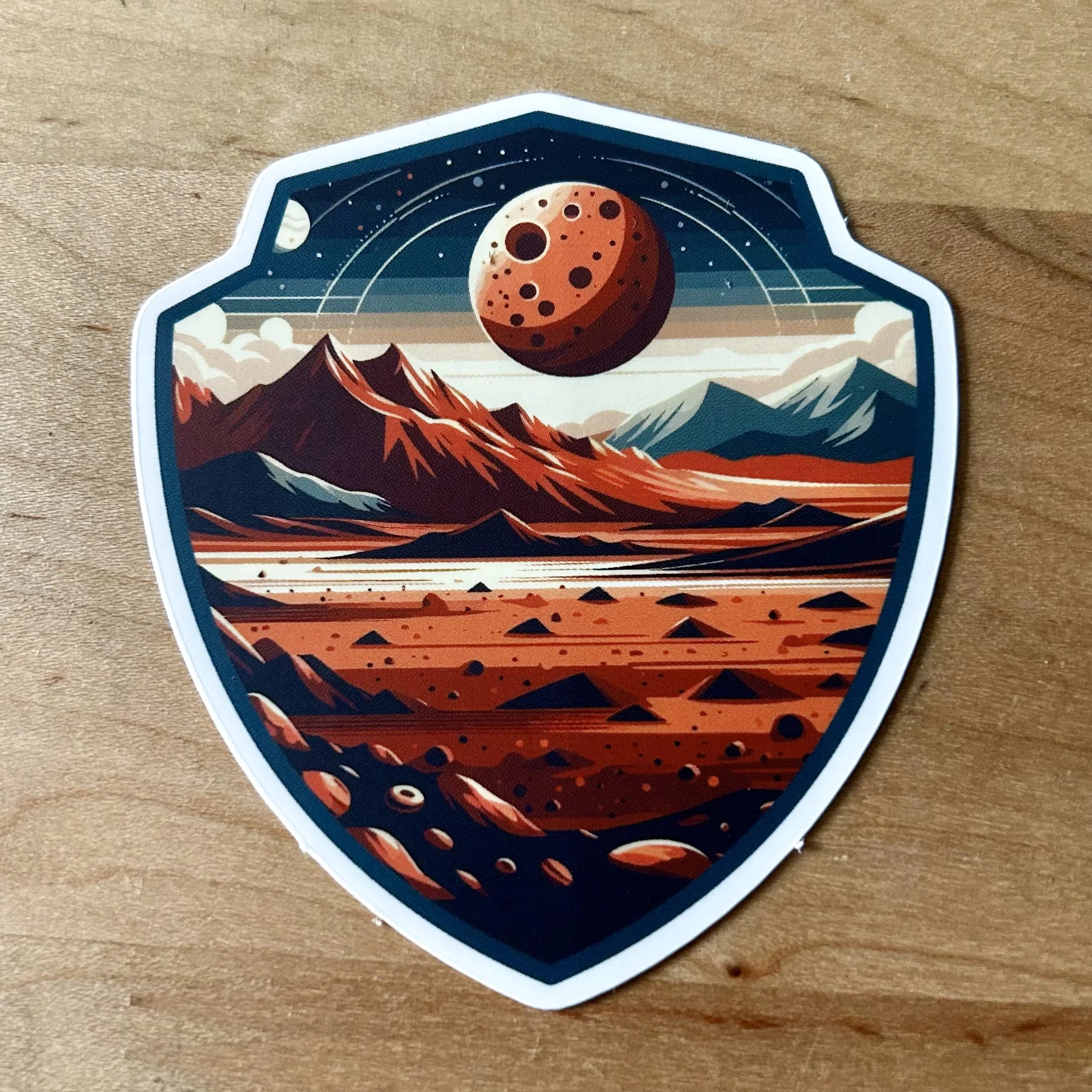 Shield-shaped sticker with red rocky landscape, mountains, and cratered planet in a starry sky