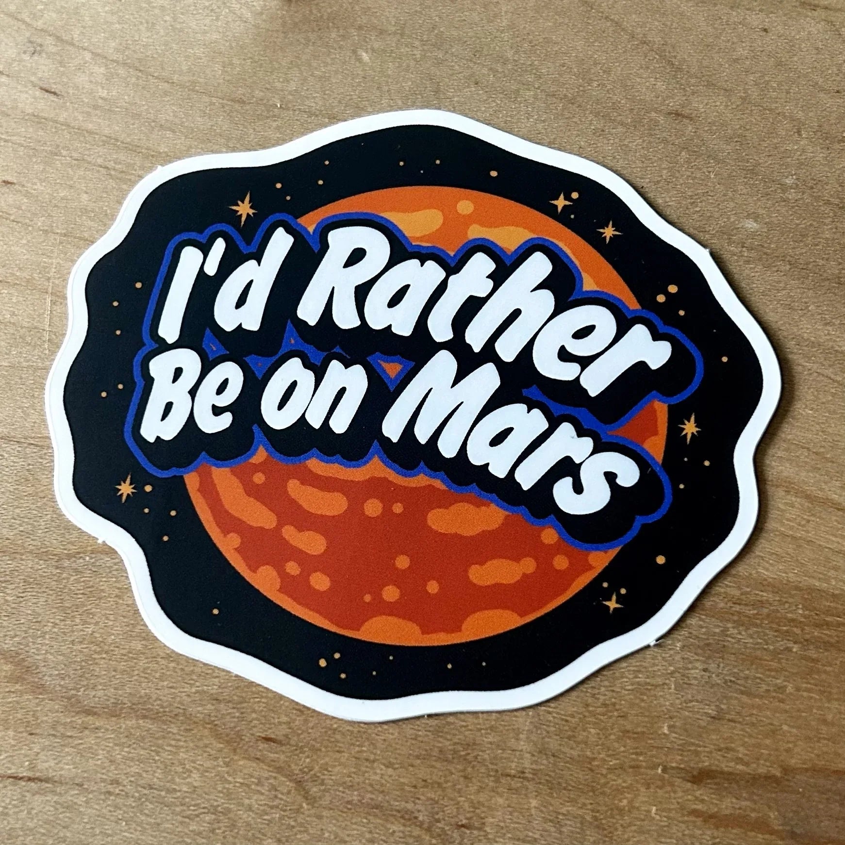 Sticker with Mars planet graphic and text I'd Rather Be on Mars on wooden surface