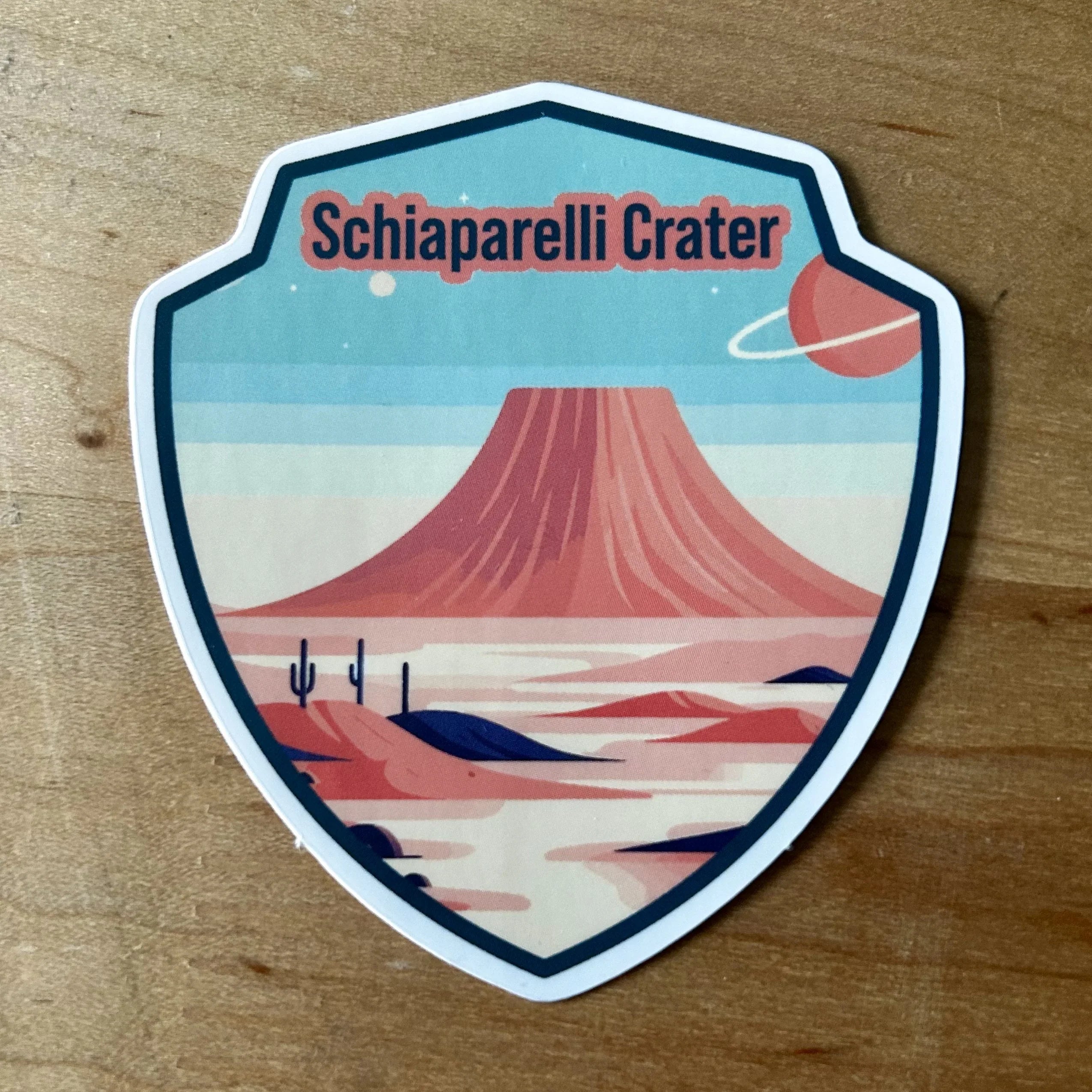Schiaparelli Crater badge with red mesa, desert landscape, cacti, and ringed planet in sky